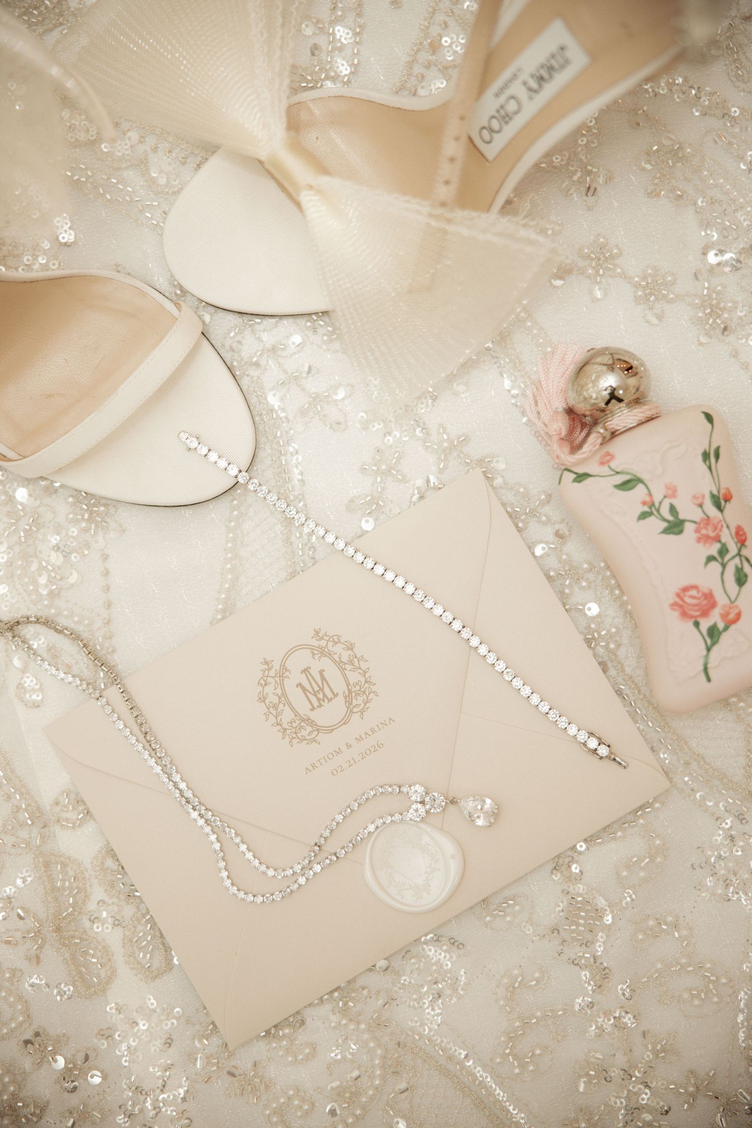 Bridal flat lay featuring white satin shoes, diamond bracelet, perfume, and monogrammed wedding invitation.