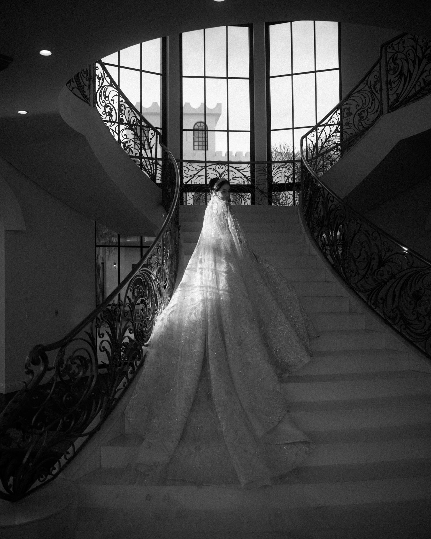 Black and white photo of bride in cathedral veil on grand double staircase in luxurious venue.