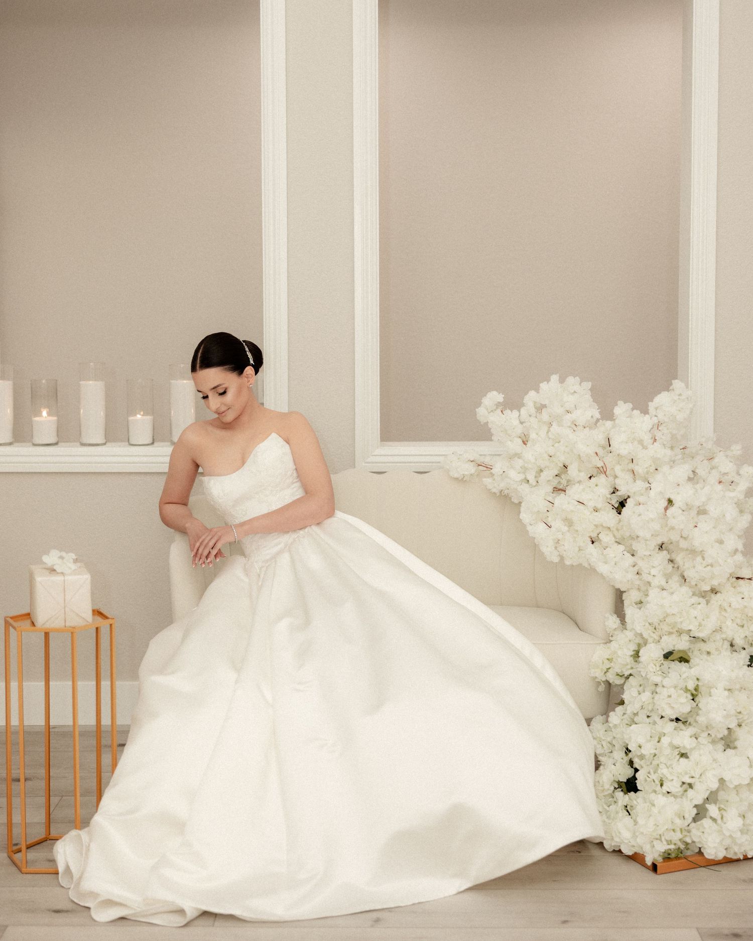 Bride in a strapless white ball gown seated near white floral arrangements and candles.