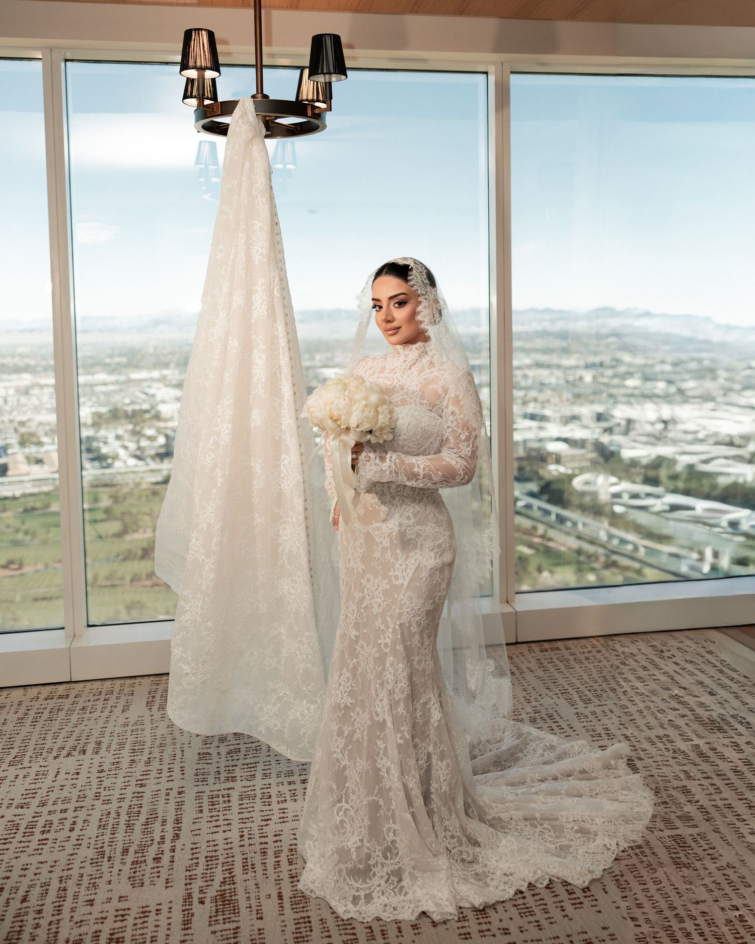 A bride in a lace wedding dress poses by floor-to-ceiling windows overlooking a cityscape.