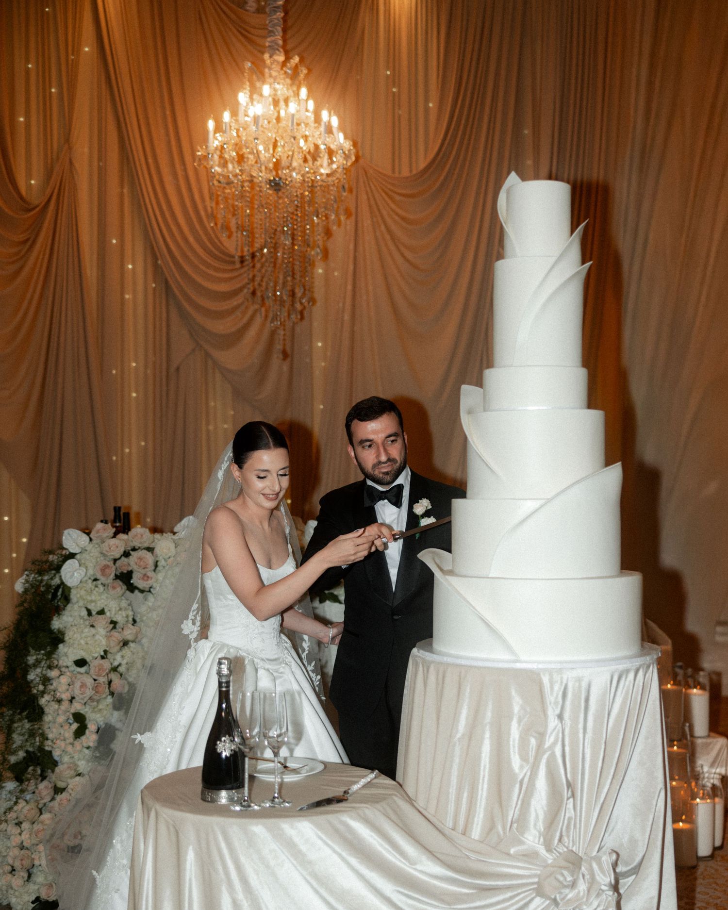 Bride and groom cutting a tall elegant white tiered wedding cake under a chandelier.