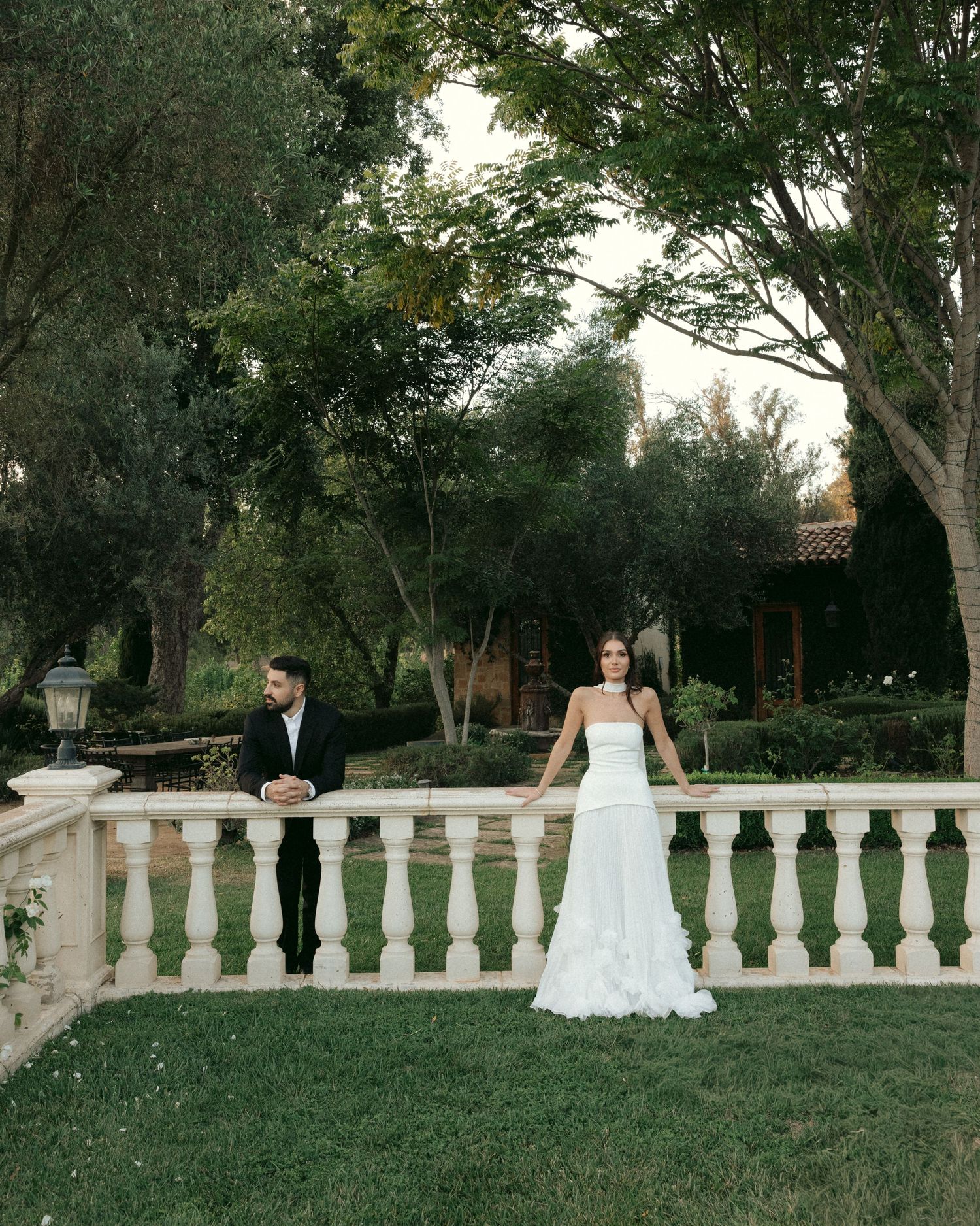 A bride in a white gown and groom in a black suit pose by a white balustrade in a lush garden setting.
