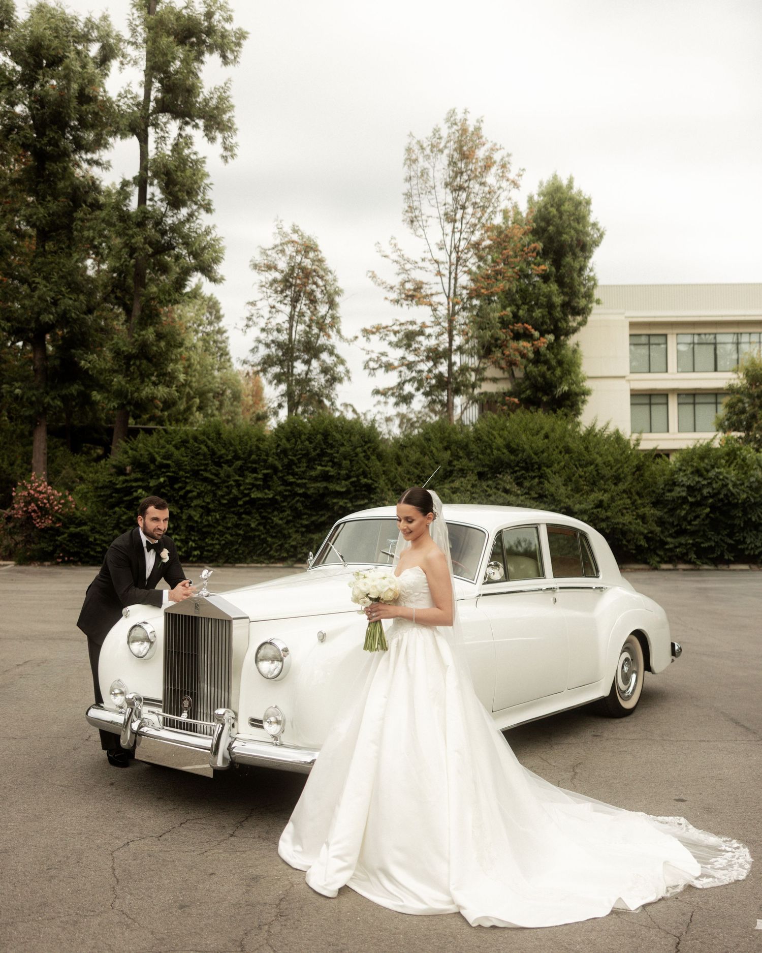 Bride and groom posing with a classic white Rolls-Royce on their wedding day outdoors.