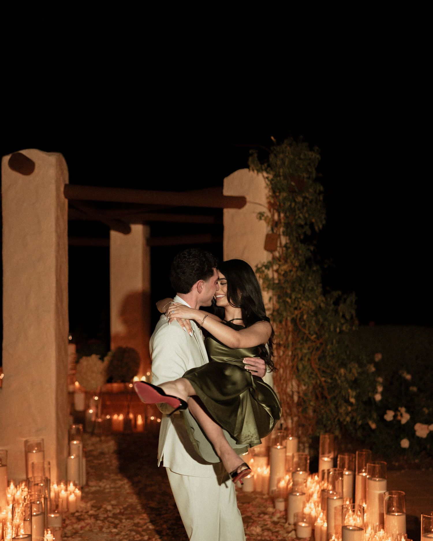 Couple embracing and kissing at night surrounded by rows of glowing candles outdoors.