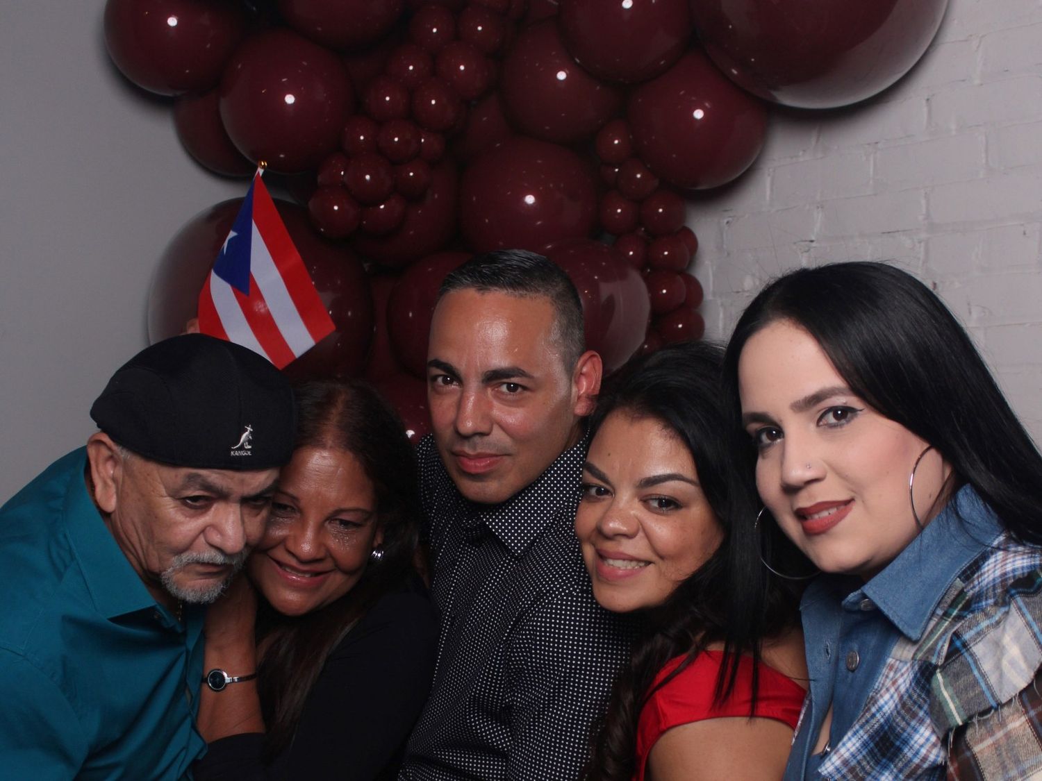 Five smiling people pose close together at a festive party in front of a wall of dark red balloons and a small Puerto Rican flag.
