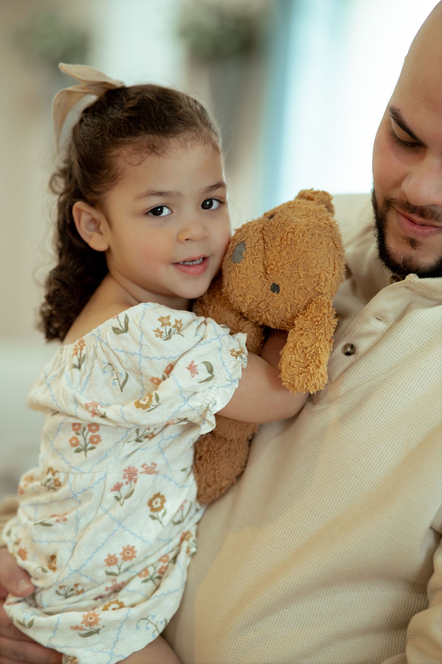 A smiling little girl in a floral dress hugs a brown teddy bear while being held by a bearded man in a cream shirt.