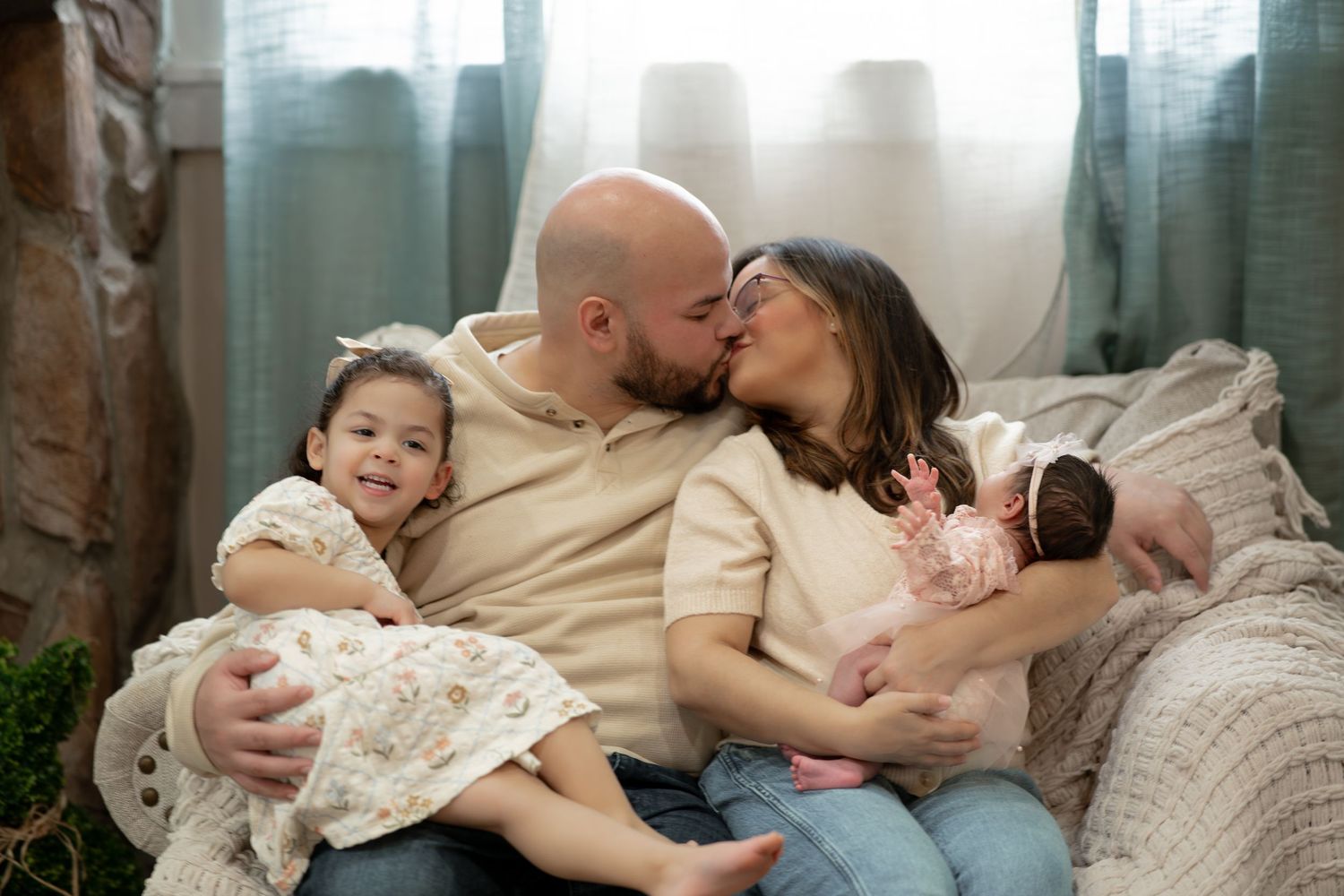 Happy family of four with parents kissing while holding newborn and toddler daughter on a soft couch.