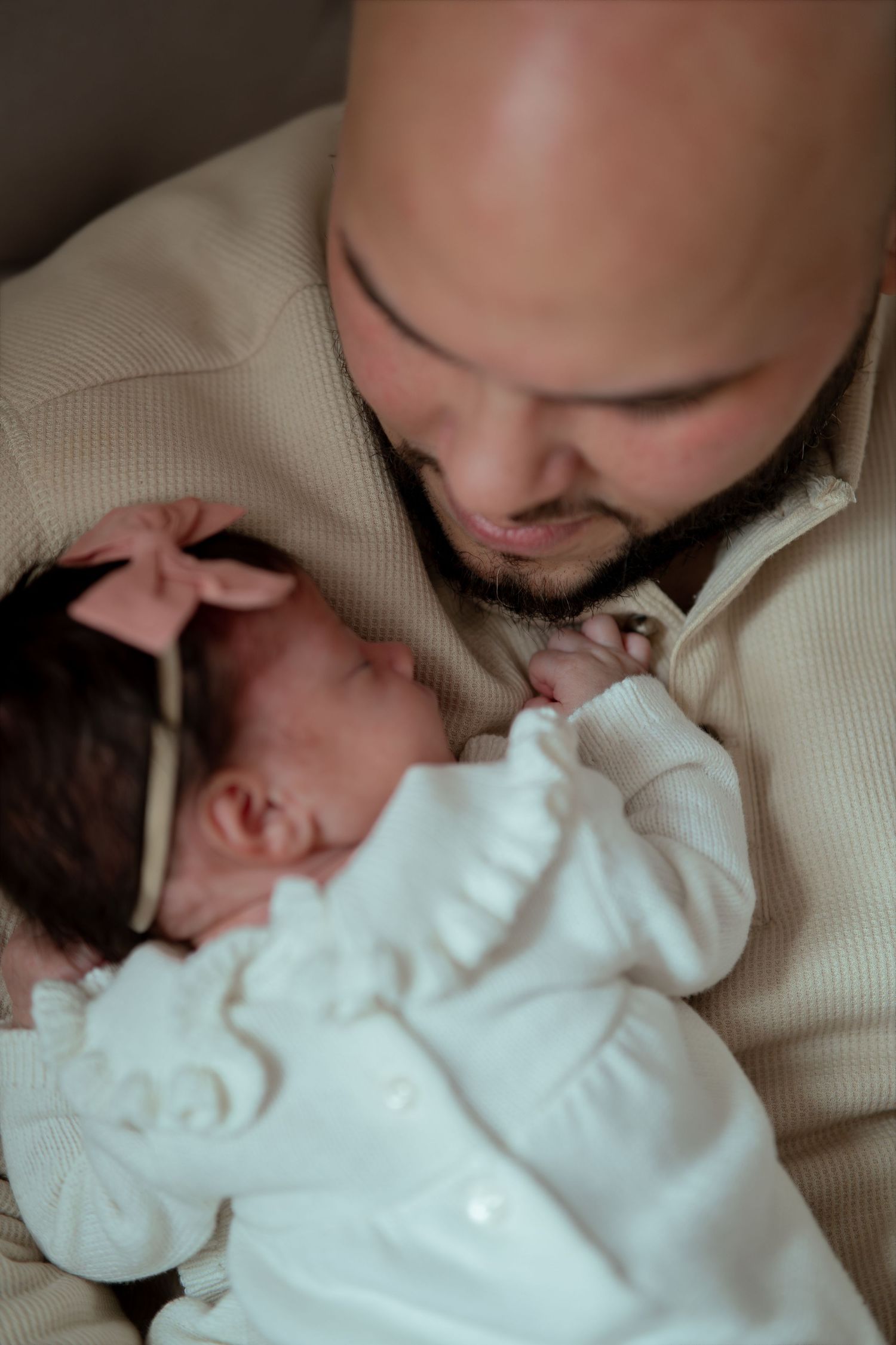 A bearded father in a cream sweater gazes lovingly at his sleeping newborn daughter dressed in white with a pink bow headband.
