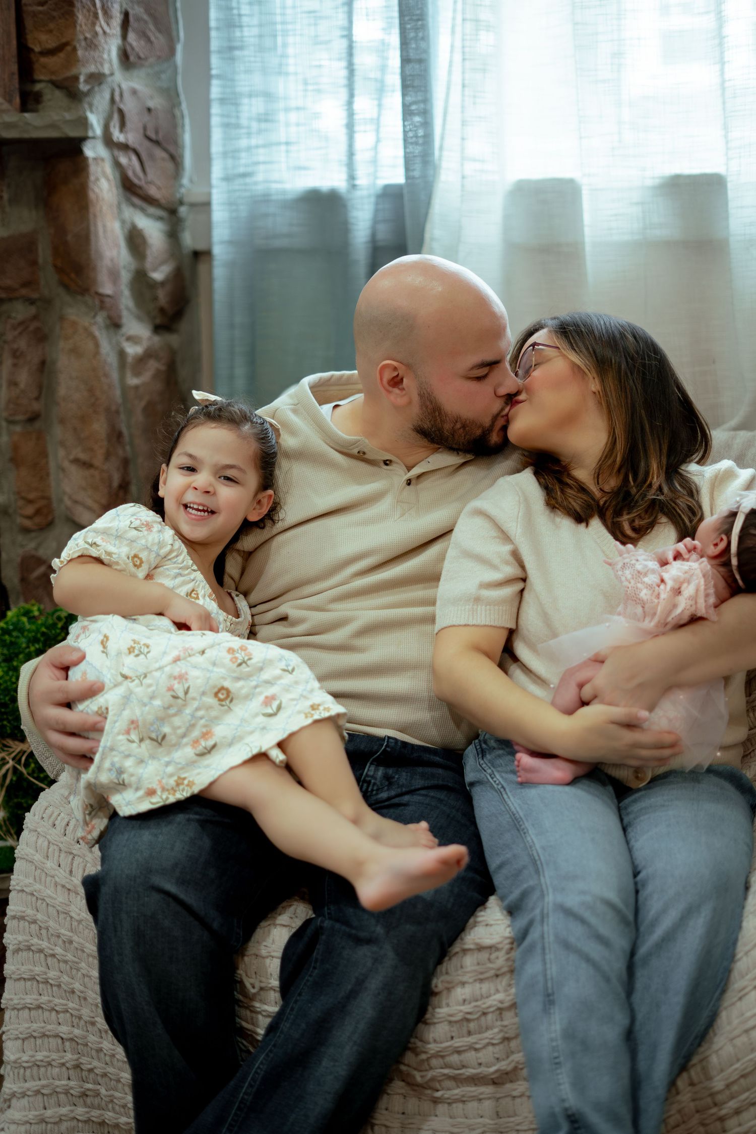 A happy family of four sits on a couch, parents kissing while dad holds toddler girl and mom cradles newborn baby.Parents kissing while hold