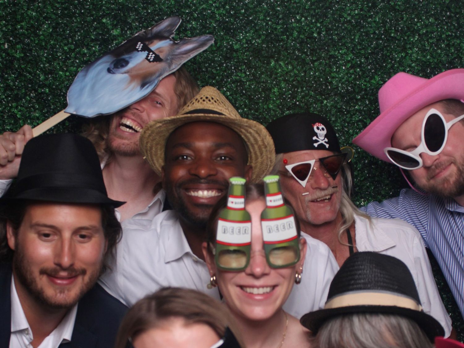 Group of smiling friends in a photo booth wearing playful hats, pirate bandana, and novelty bottle glasses against a green hedge wall.