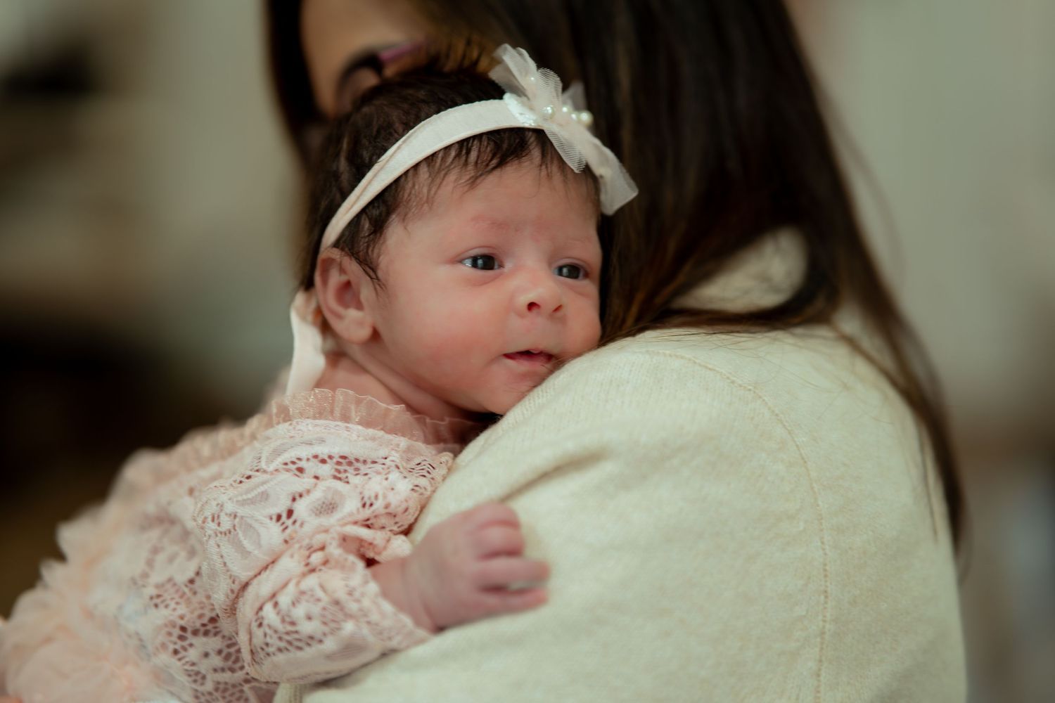 A newborn baby in a white bow headband and lace outfit rests against a woman's shoulder while looking calmly at the camera.