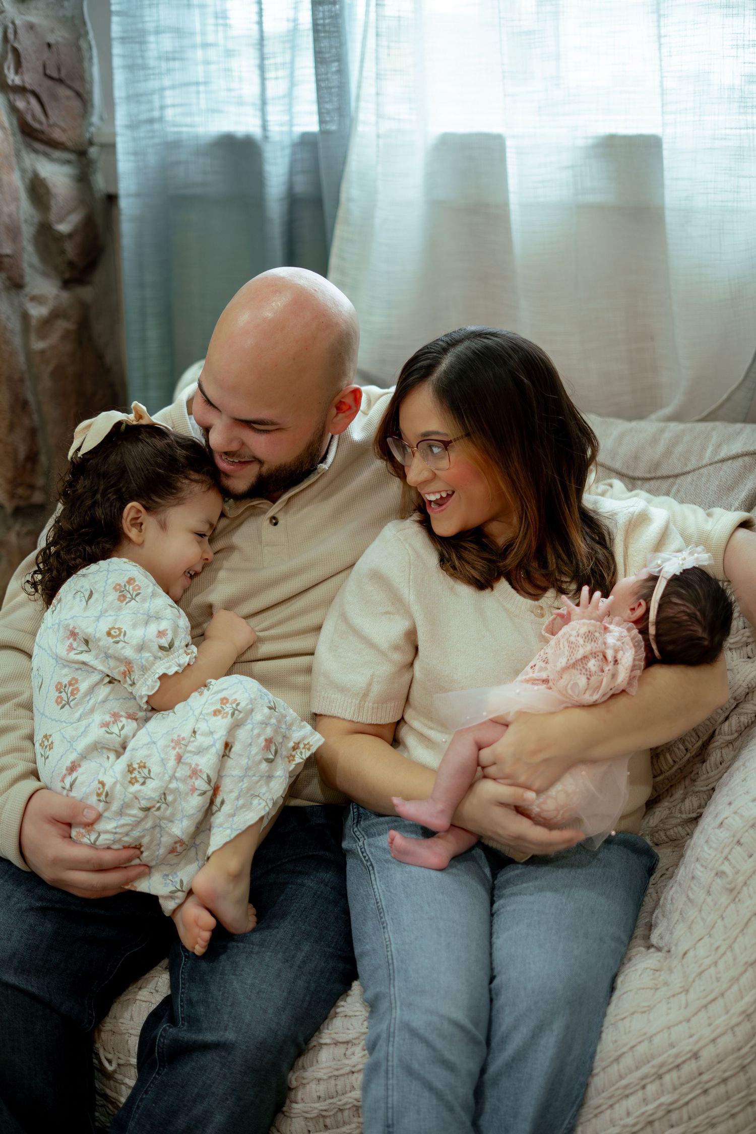 Smiling parents sit on a couch with a toddler girl and a sleeping newborn baby in pink, sharing a joyful family moment.