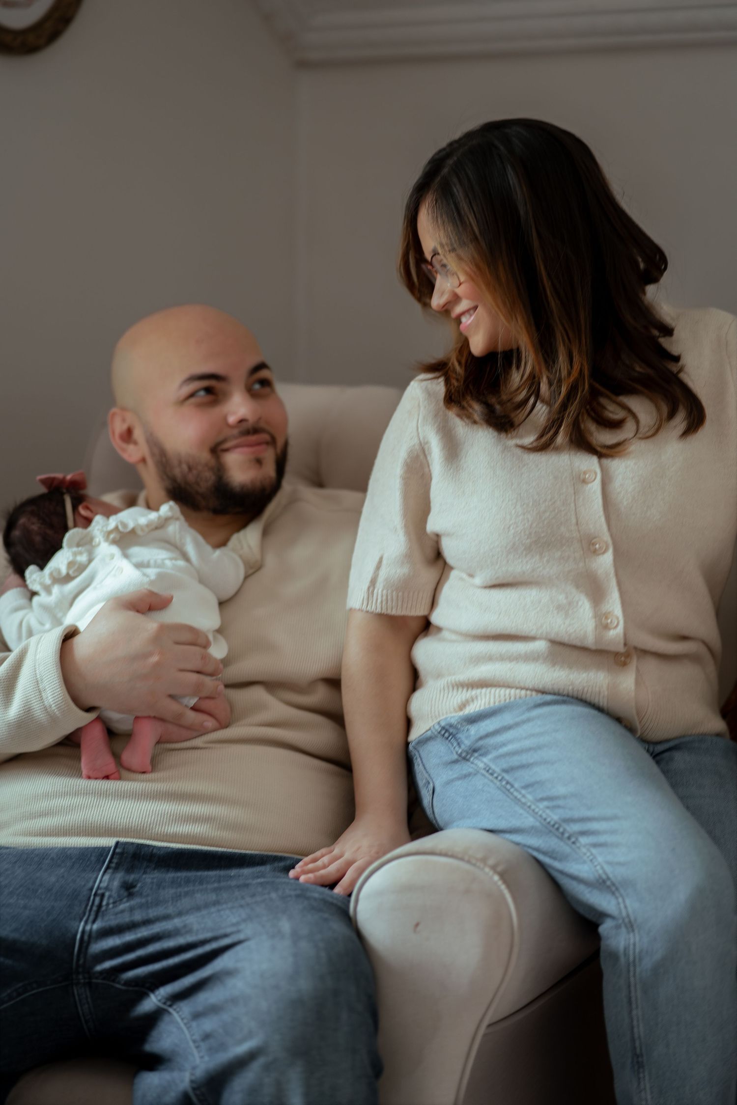 A smiling bearded man holds a newborn baby on a cream sofa while a woman in a knit cardigan sits on the armrest and looks down at them.