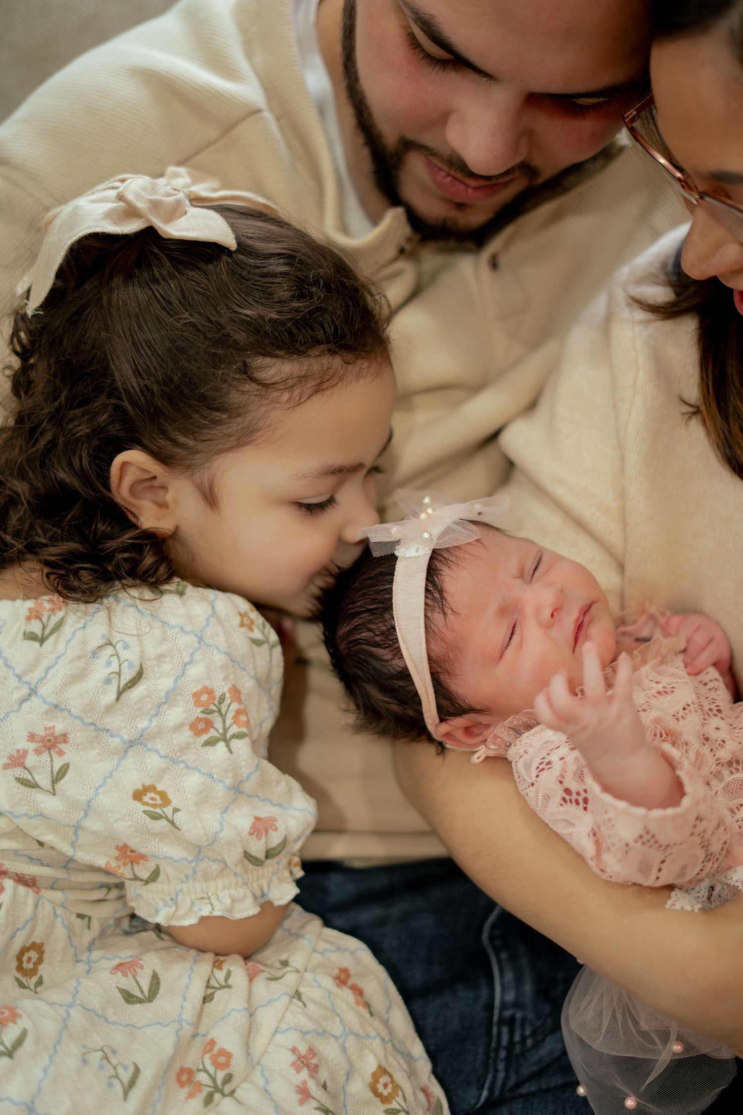 A young girl in a floral dress and bow gently kisses a sleeping newborn baby while her parents hold them close.