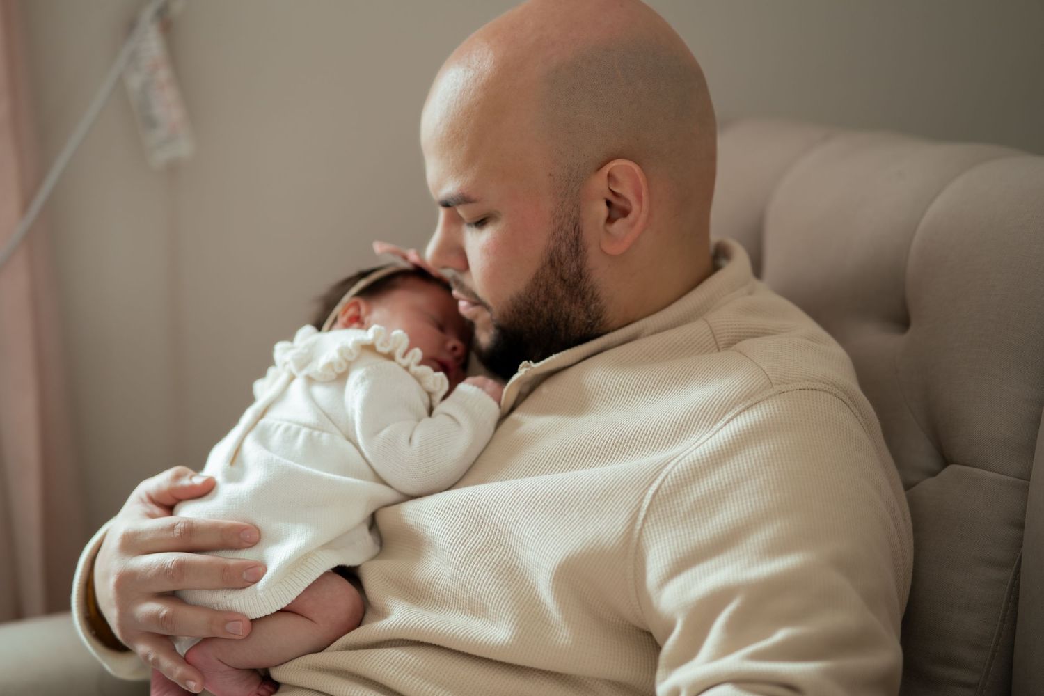 Bald bearded father in a cream sweater gently cradles a sleeping newborn baby in white against his chest while seated on a tufted couch.