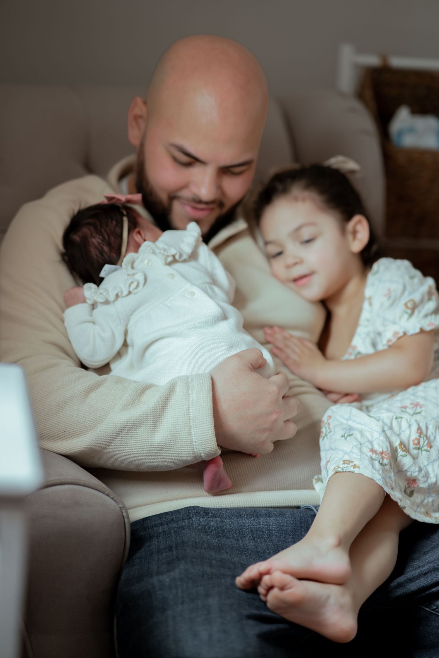 Bald father smiles holding newborn baby while toddler daughter leans in adoringly on a cozy armchair.