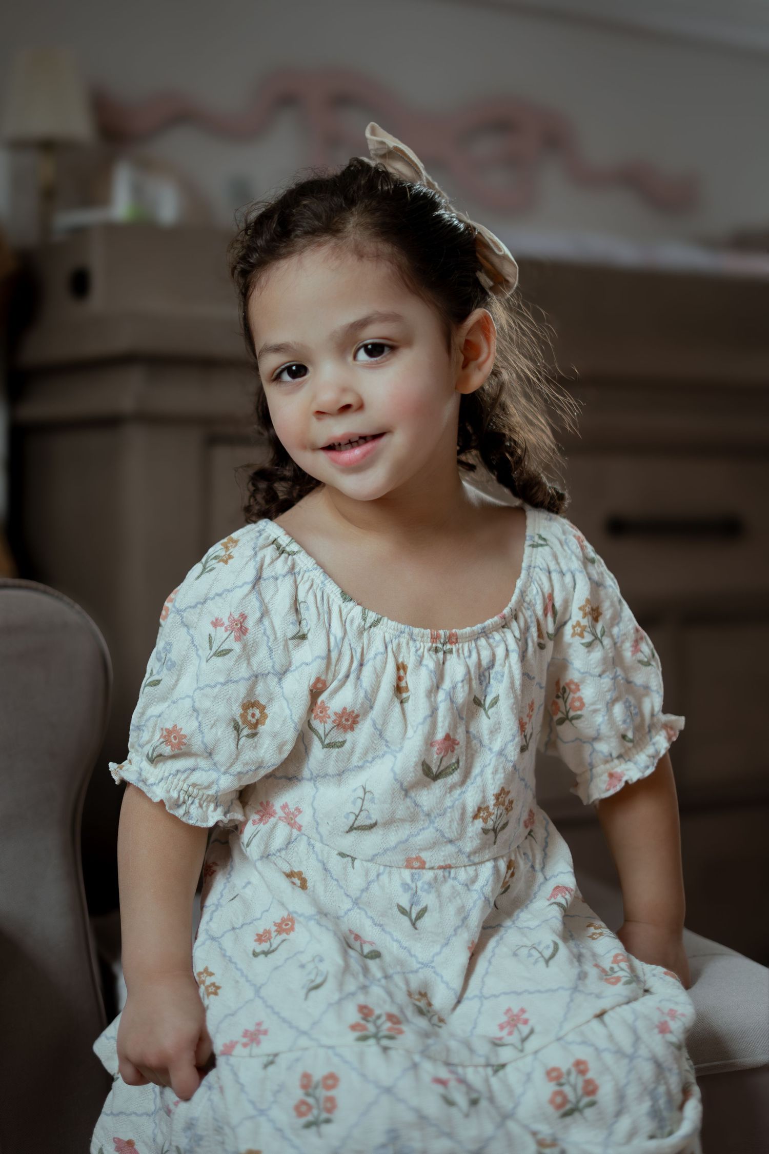 A smiling toddler girl with curly hair and a bow sits on a chair in a floral puff-sleeve dress in a cozy, softly lit room.