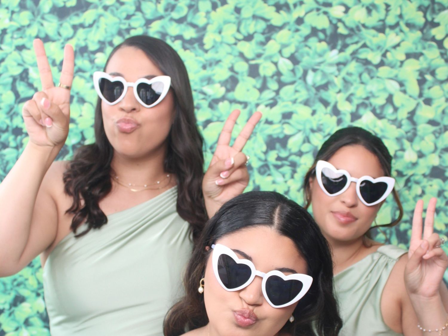 Three women in sage green dresses pose with peace signs and pouty lips in white heart-shaped sunglasses before a green leafy photo booth bac