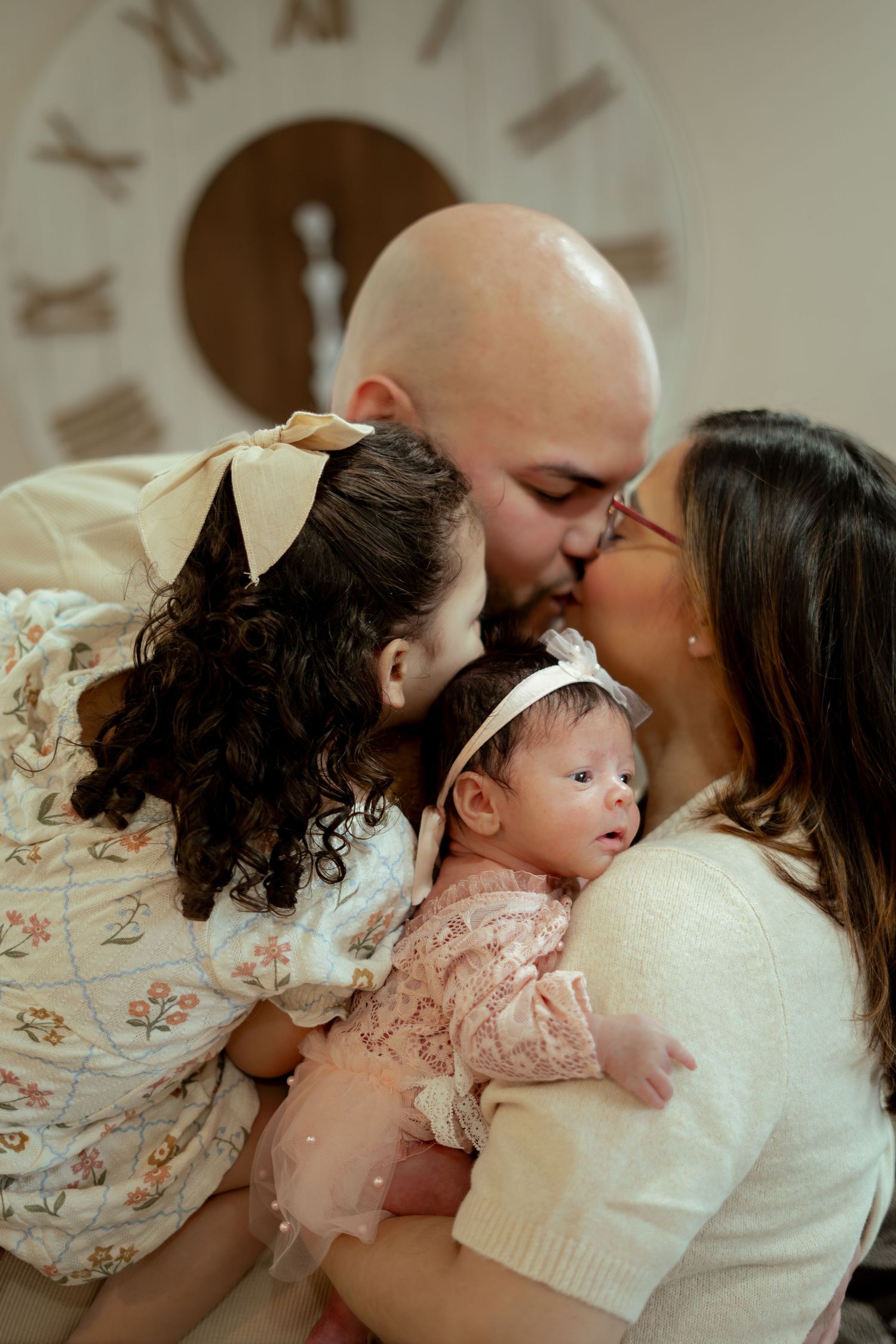 A family shares a tender kiss as parents cuddle a newborn baby in a pink outfit and a curly-haired girl with a bow near a large wall clock.