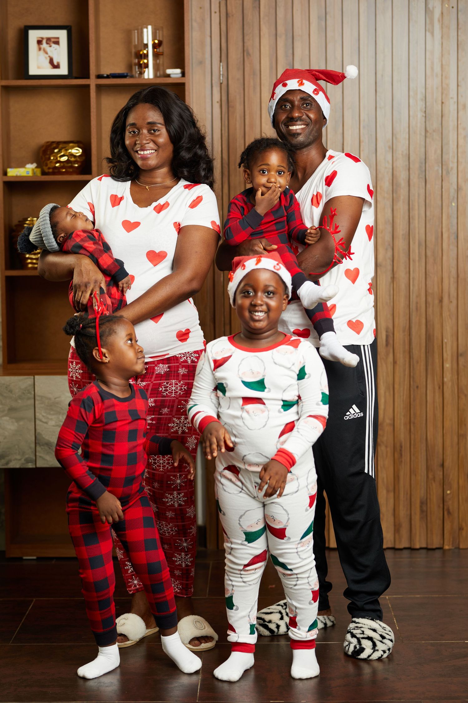 Family wearing matching holiday pajamas with heart patterns pose in front of wooden wall.