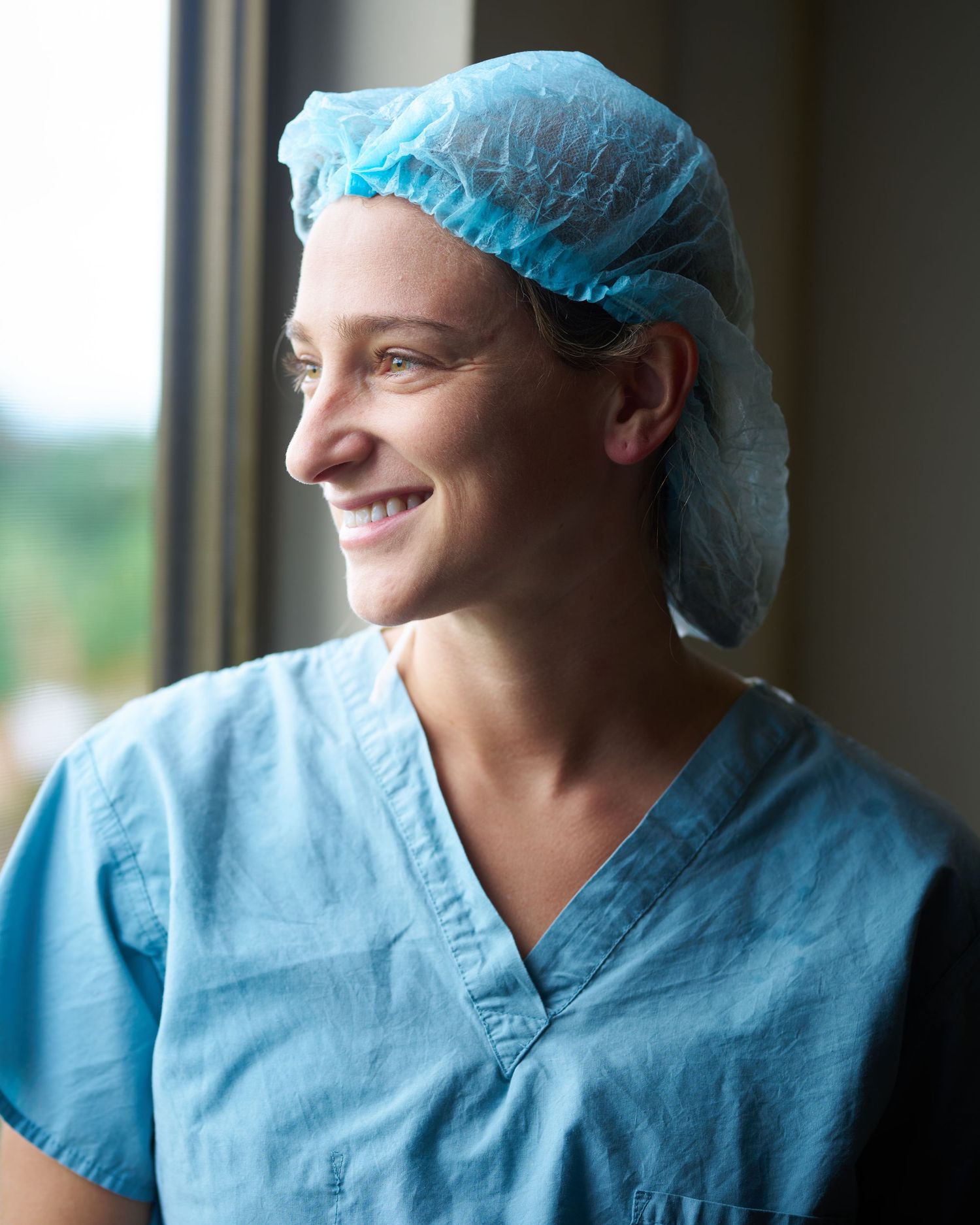 Healthcare worker in blue scrubs and cap looking thoughtfully through hospital window.