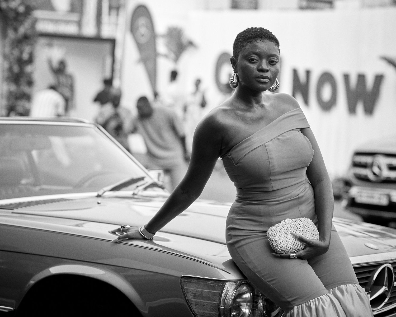 Black and white photo of an elegant woman in a fitted dress leaning on a vintage Mercedes-Benz car outdoors.