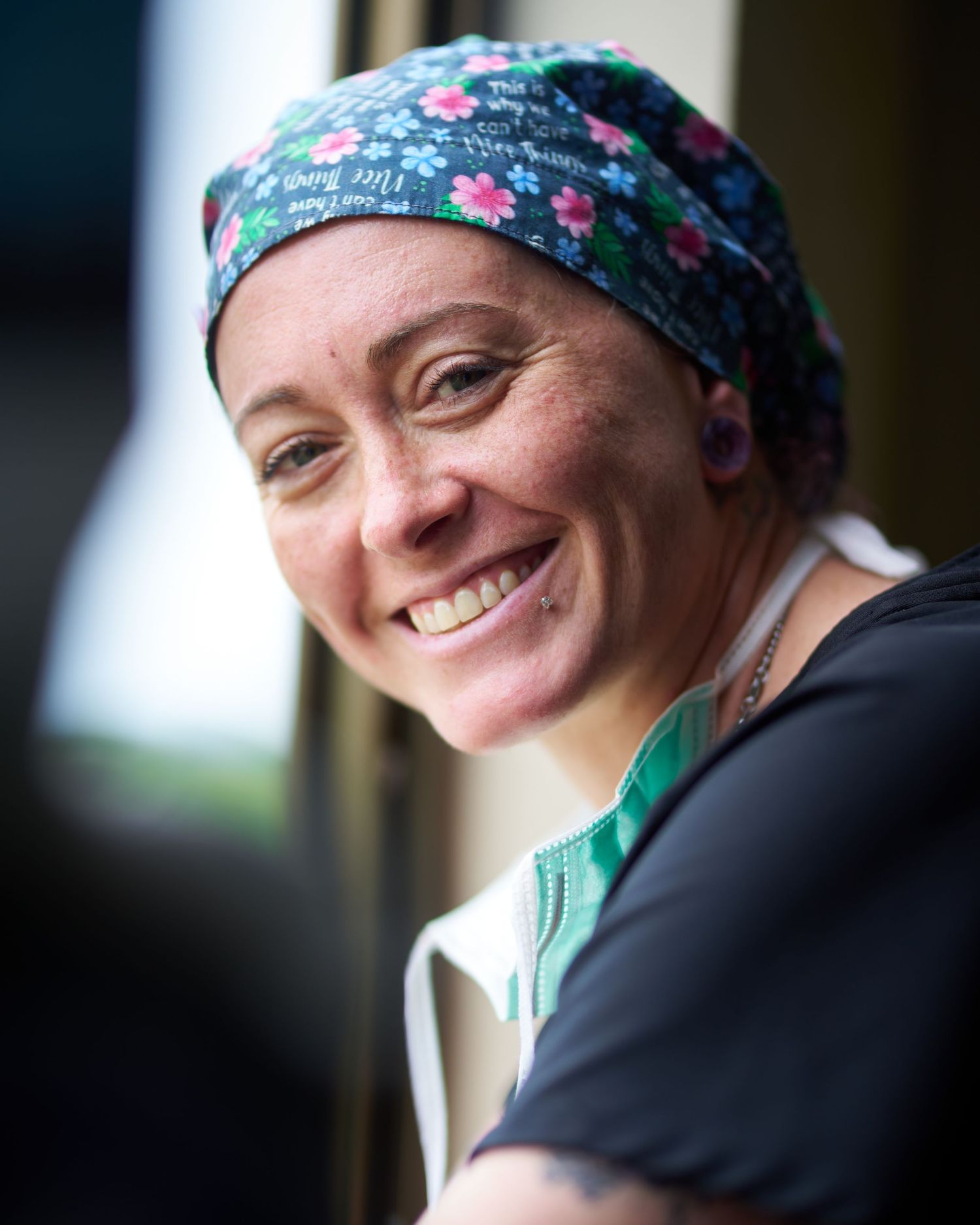 Healthcare worker wearing floral scrub cap smiles warmly in natural lighting.