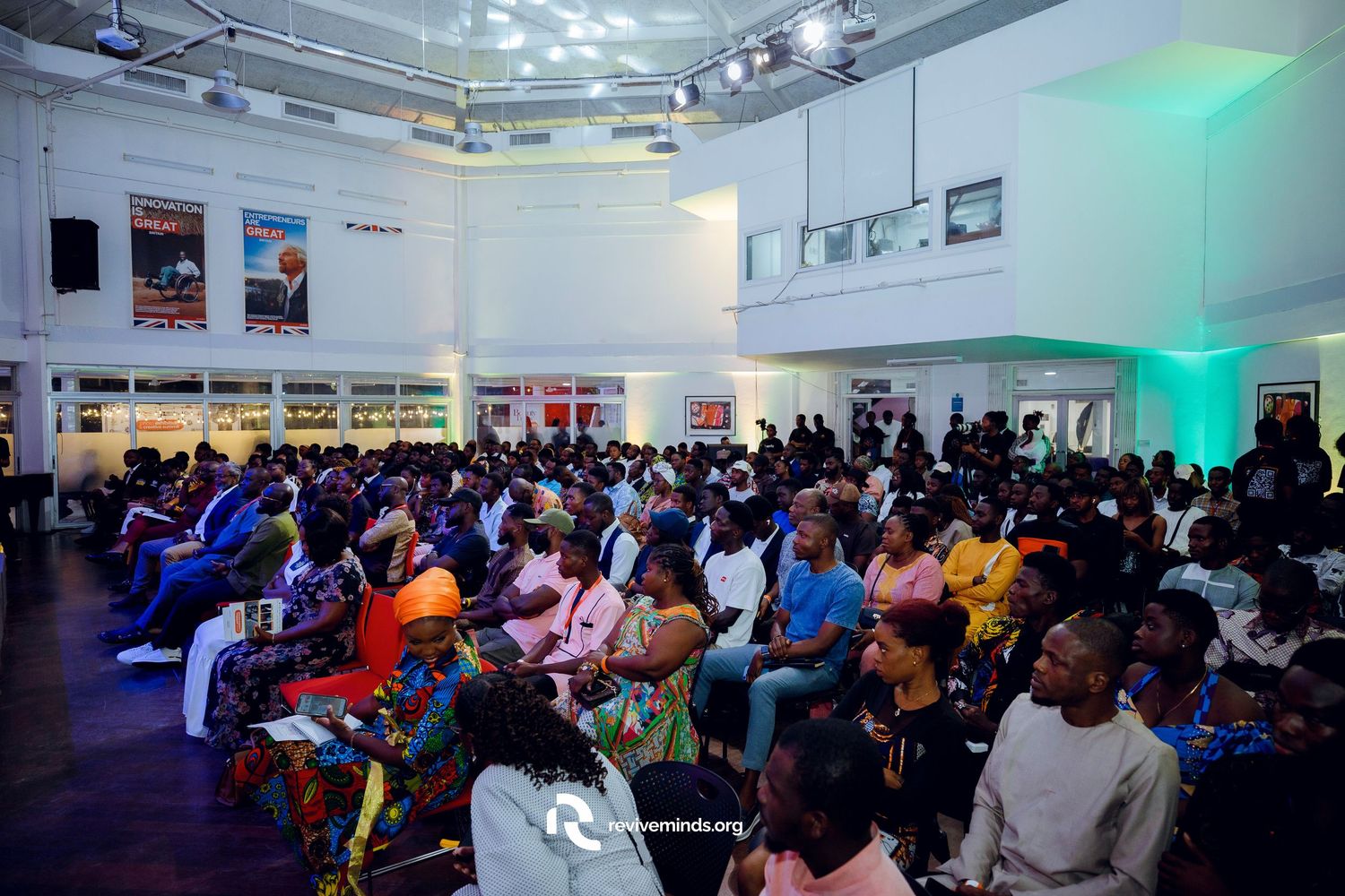 Wide shot of large audience seated in modern event space with blue lighting.