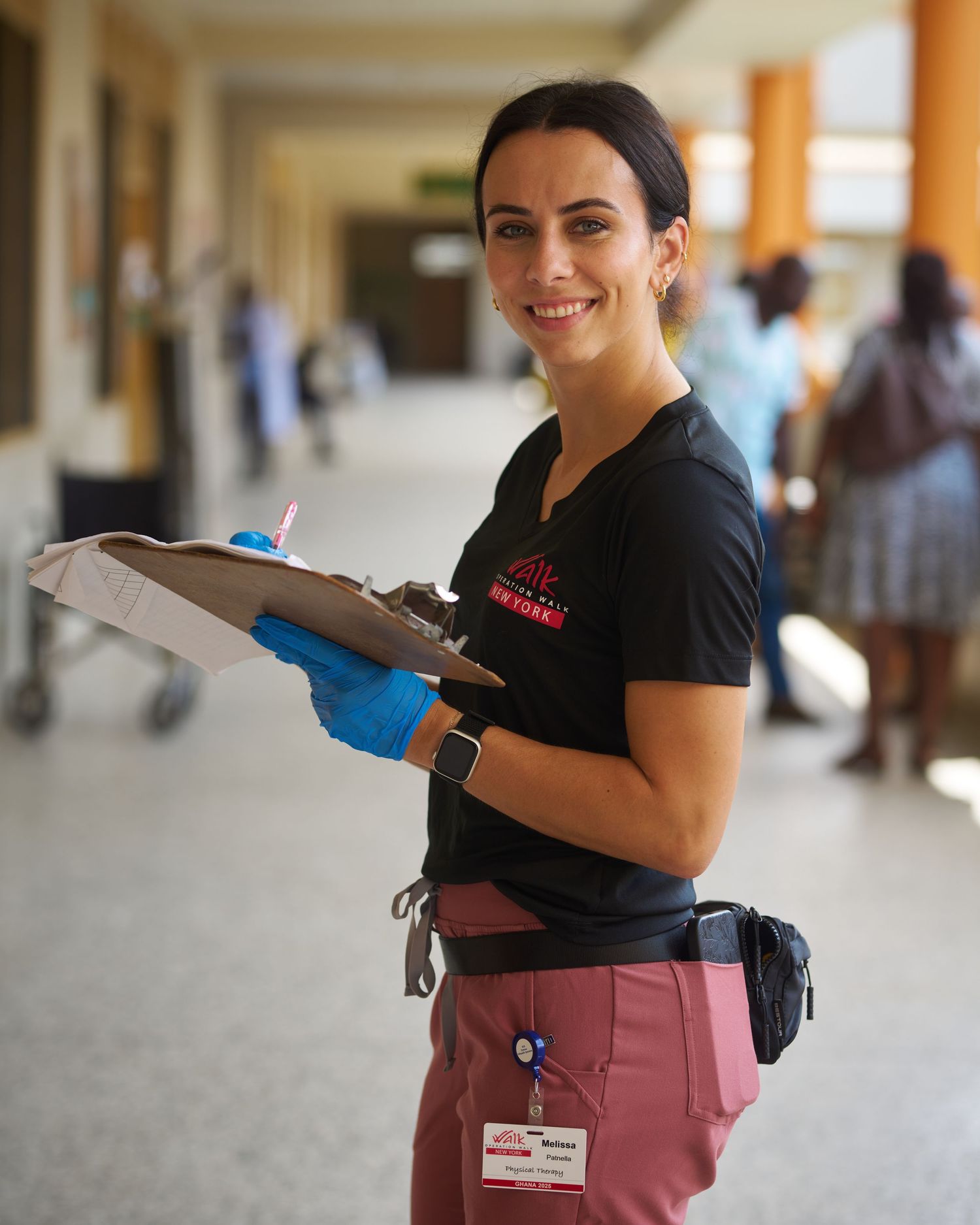 Hospital staff member in uniform carrying clipboard through medical facility hallway.