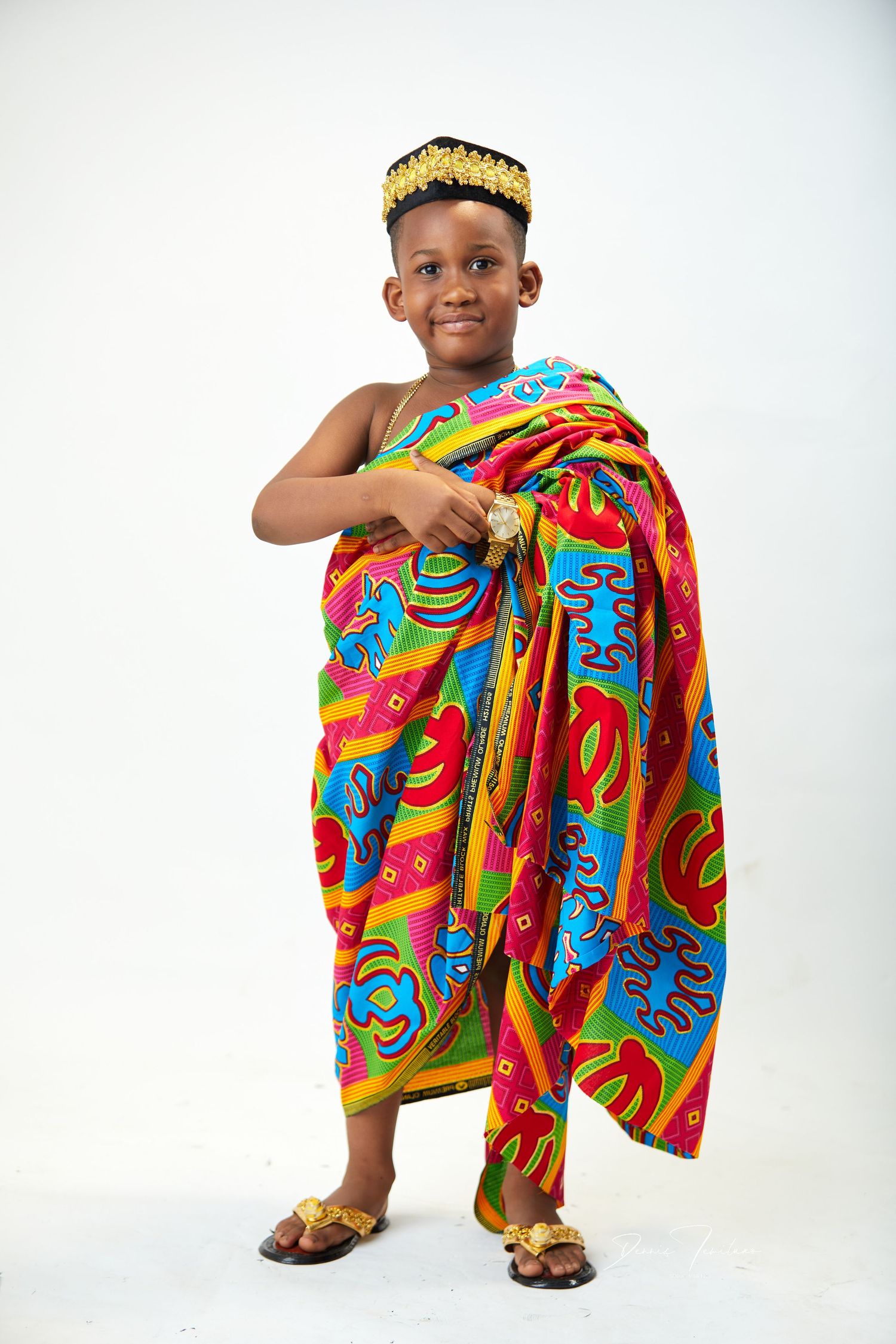 Young child wearing colorful traditional African attire and crown poses against white background.