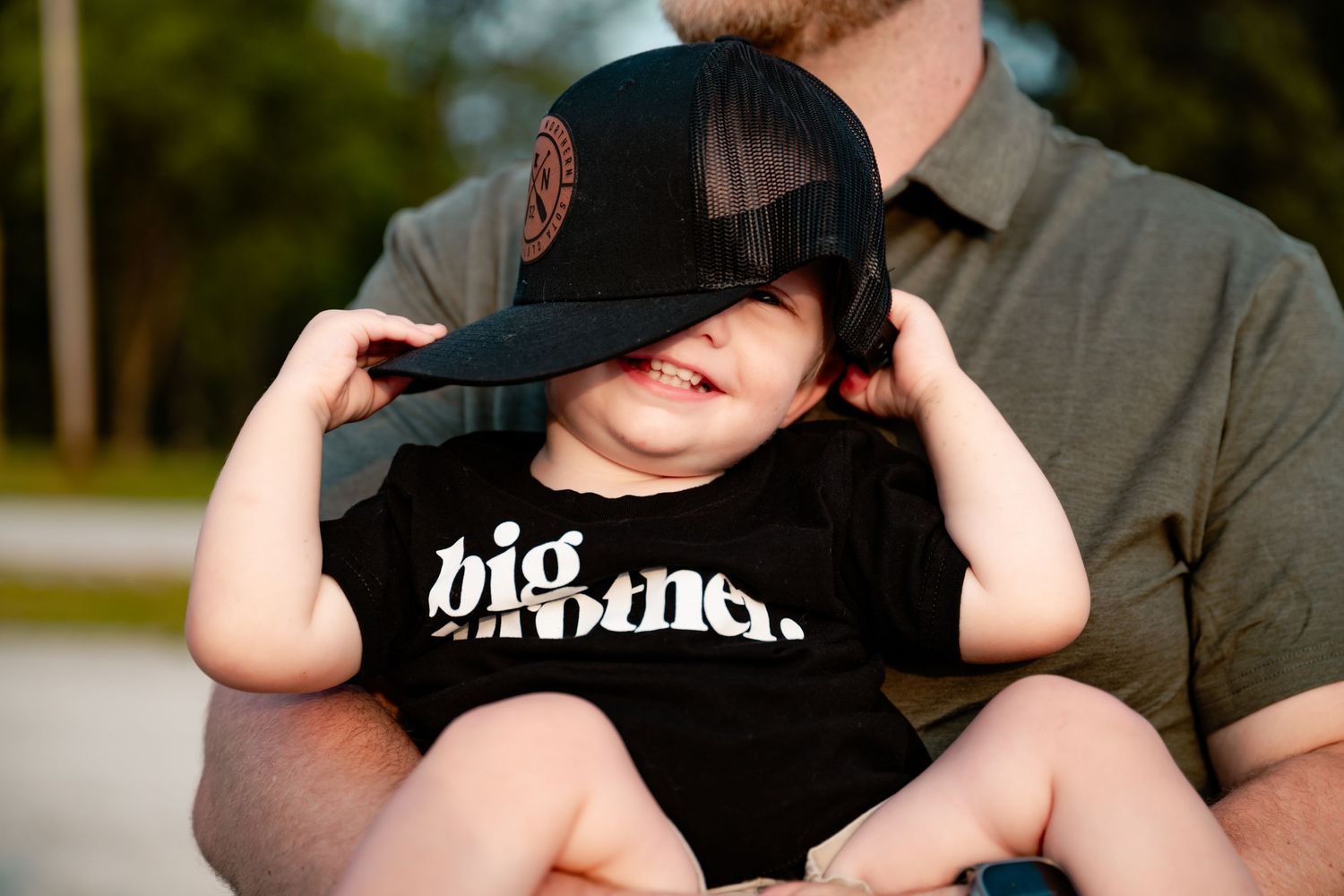 Small child wearing oversized baseball cap and big brother t-shirt being held by parent.