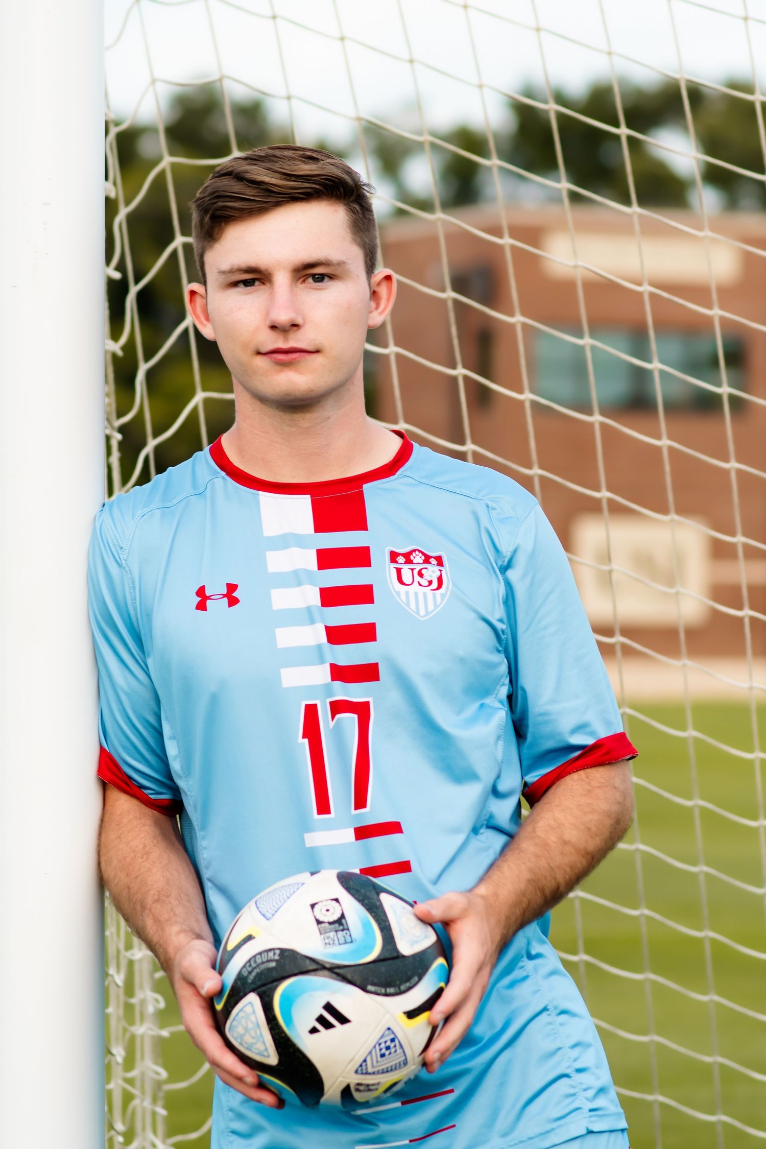 A soccer goalkeeper in a light blue uniform with red accents poses with a ball beside the goal net.