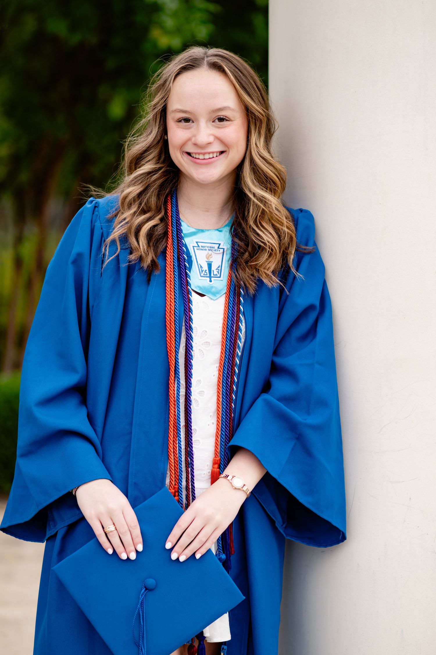 Graduate in blue academic regalia smiling while holding cap against white column.