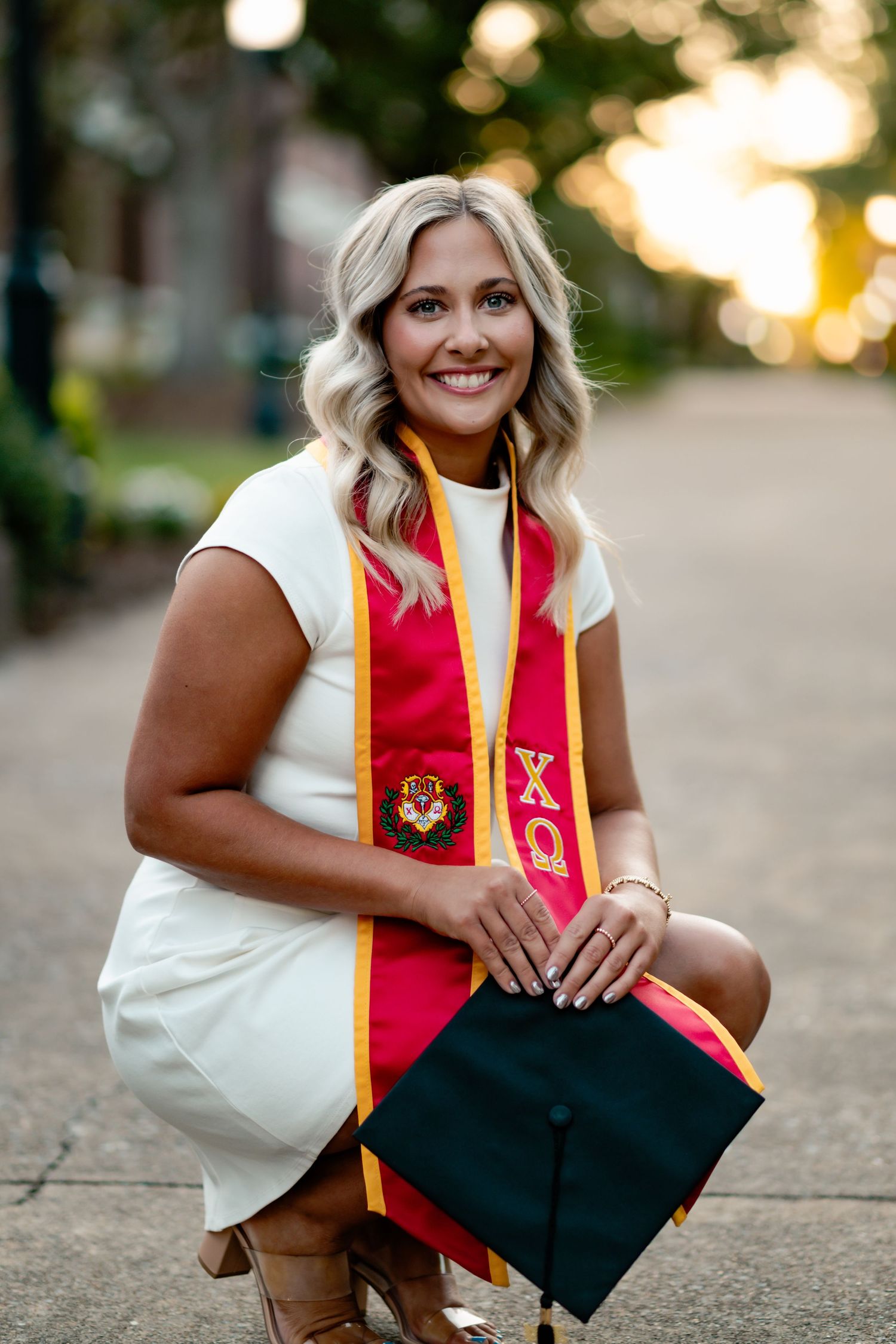 Graduate in white dress celebrates with red and gold graduation stoles on campus.