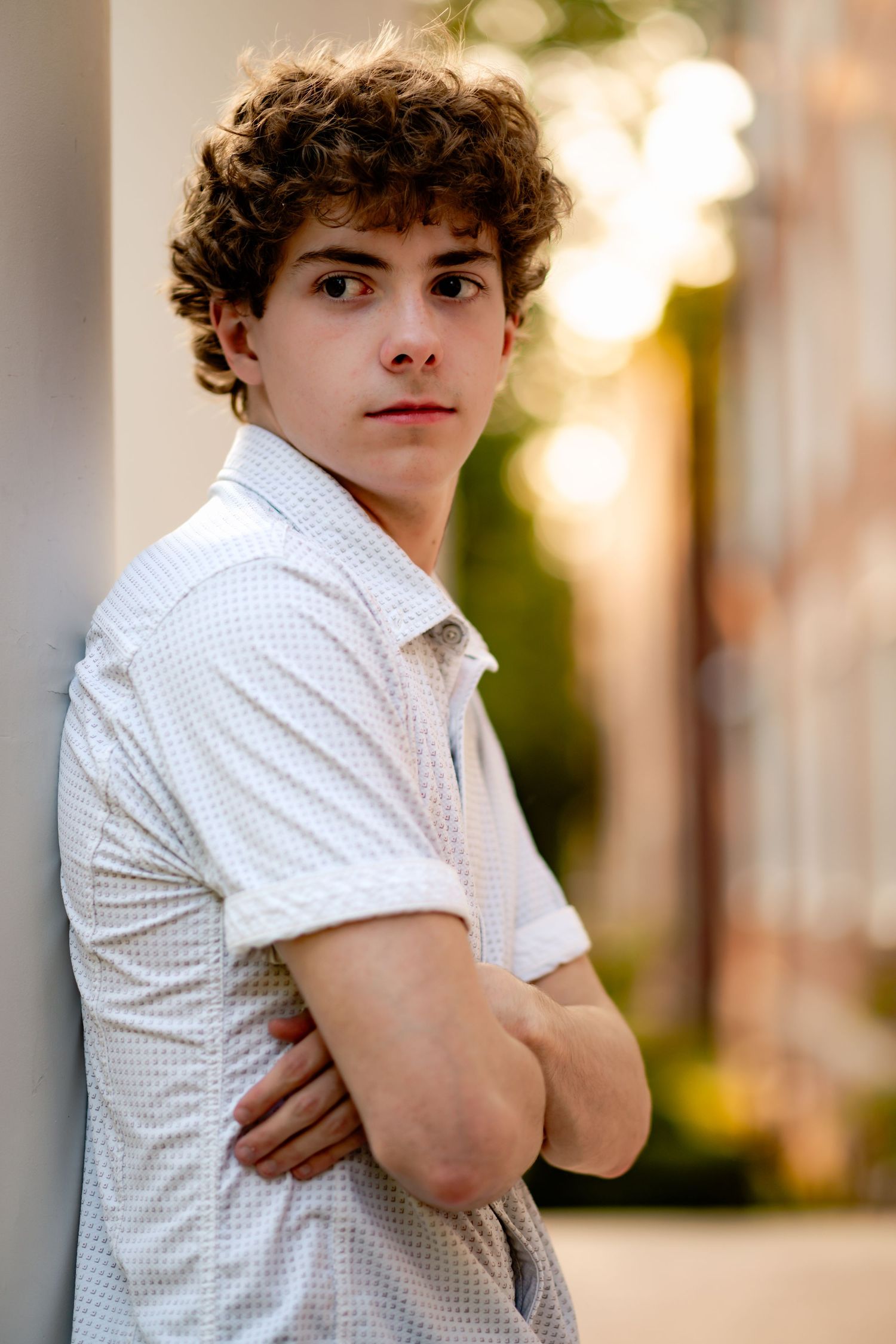 Portrait photo outdoors of a young person in a white polka dot shirt standing with arms crossed in warm lighting.