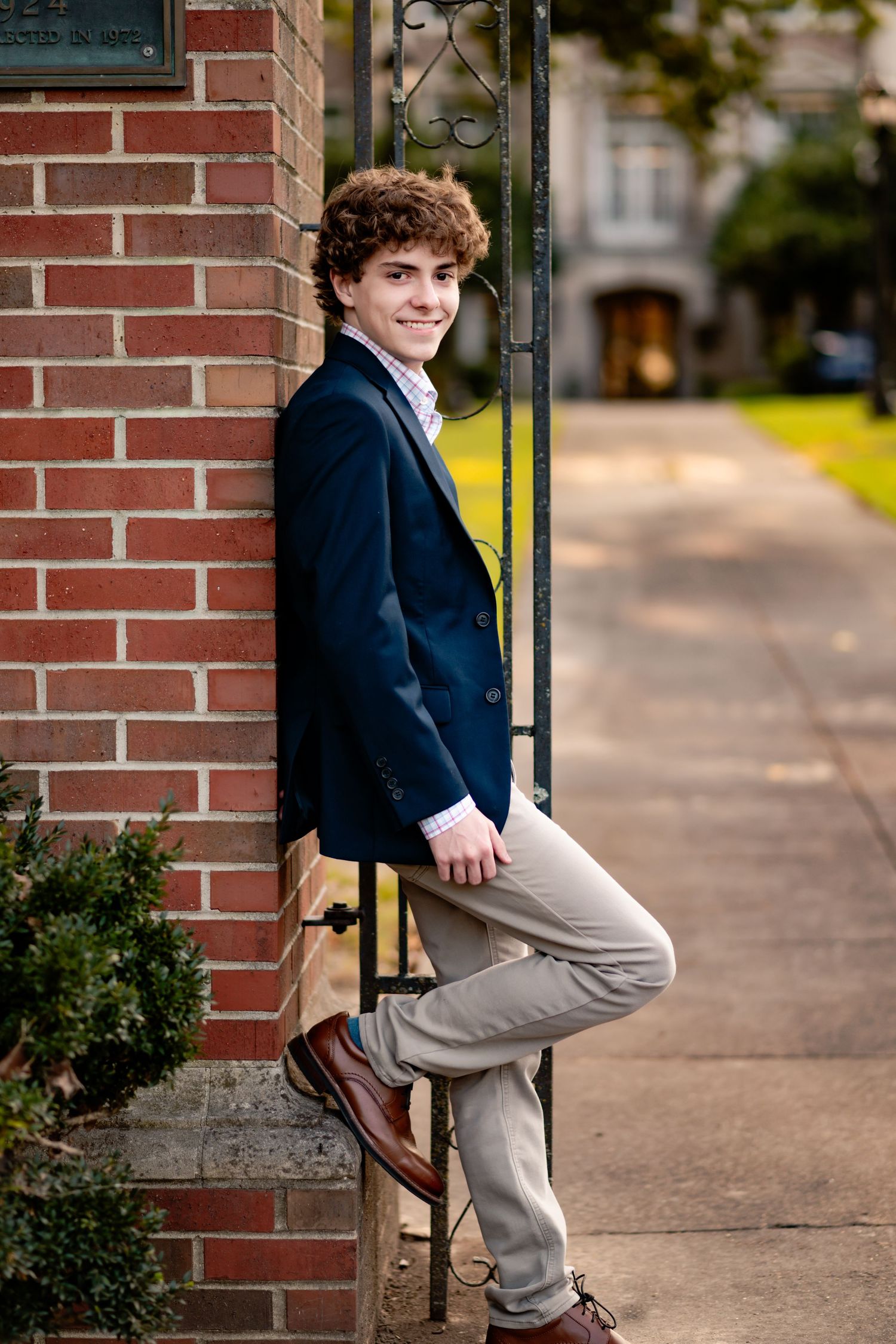 Student in navy blazer and khaki pants poses by brick wall and column during outdoor portrait session.