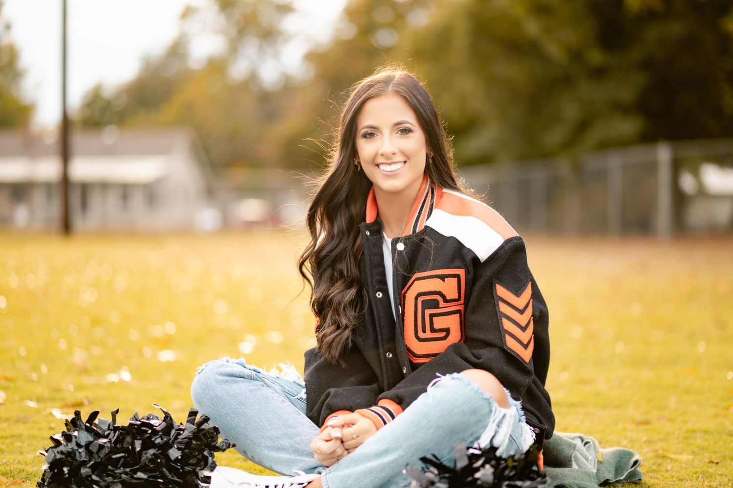 Senior student in high school jersey poses with black pom poms in grassy field on a fall day.