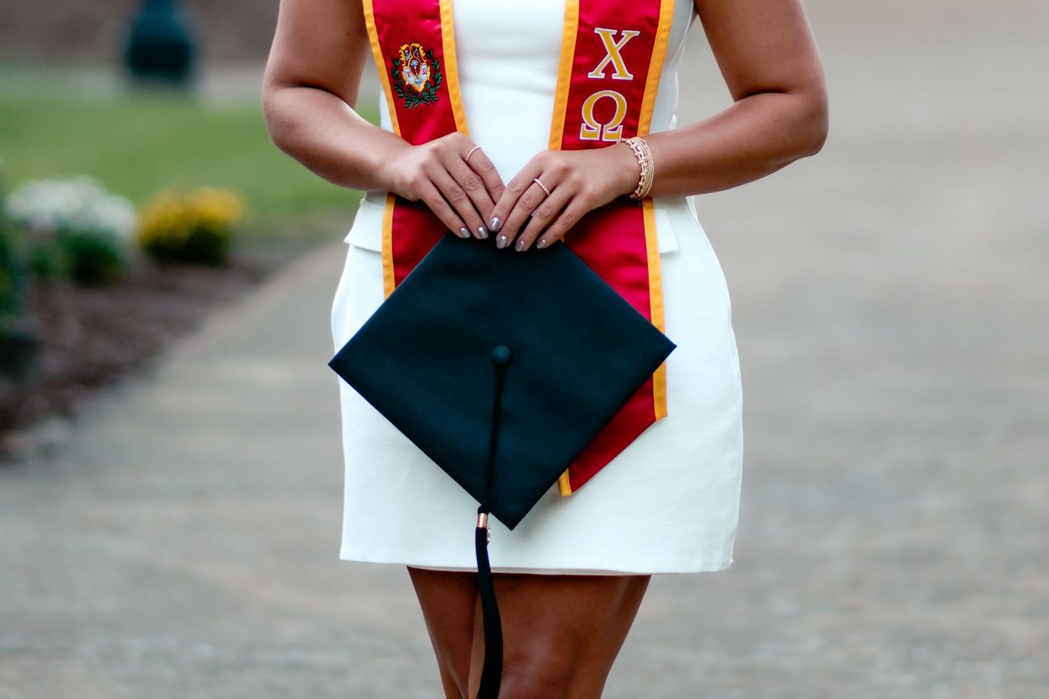 Graduate wearing red and gold honor cords poses with diploma against campus backdrop.