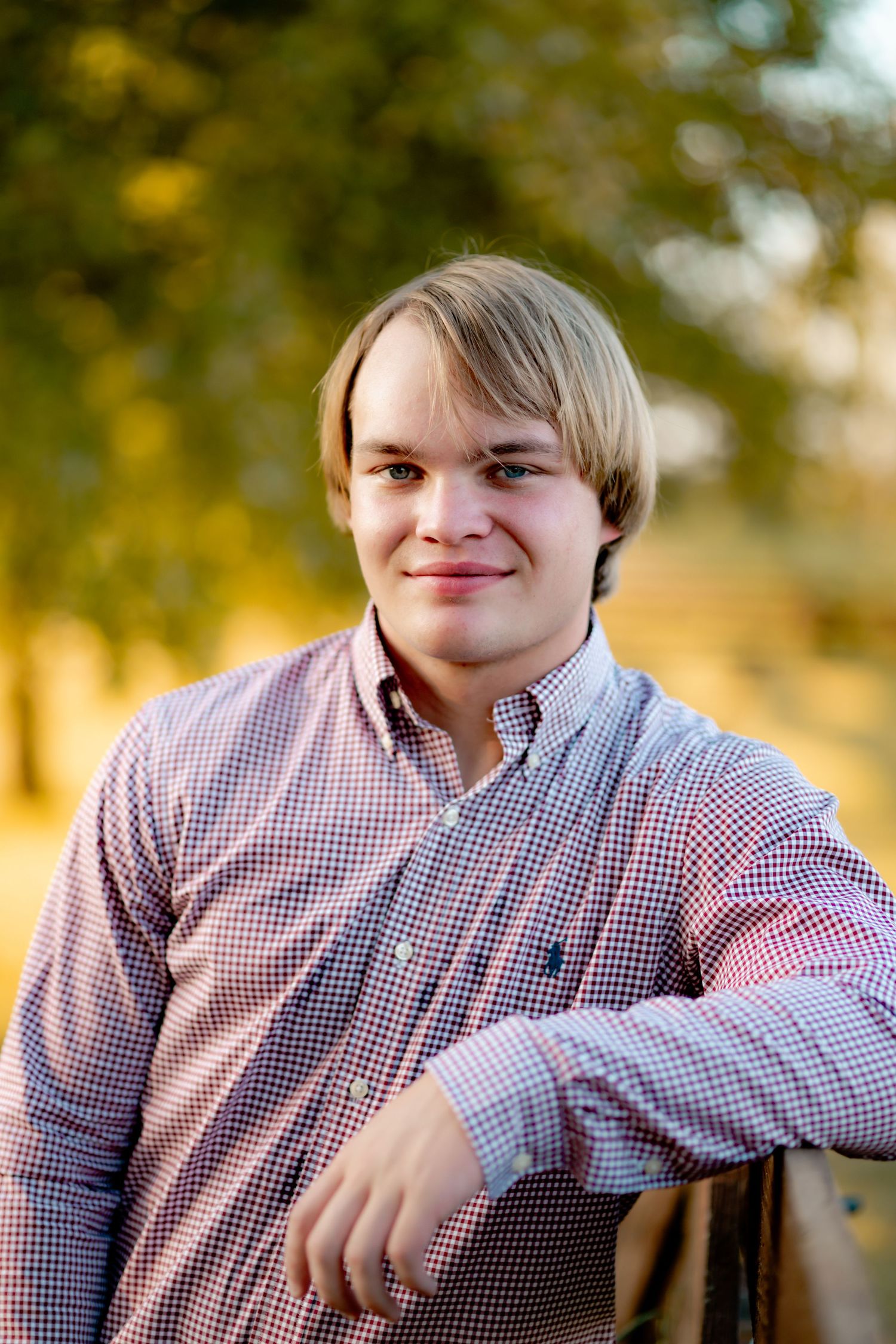 Portrait of a young person wearing a red and white checkered button-down shirt outdoors with autumn foliage in background.