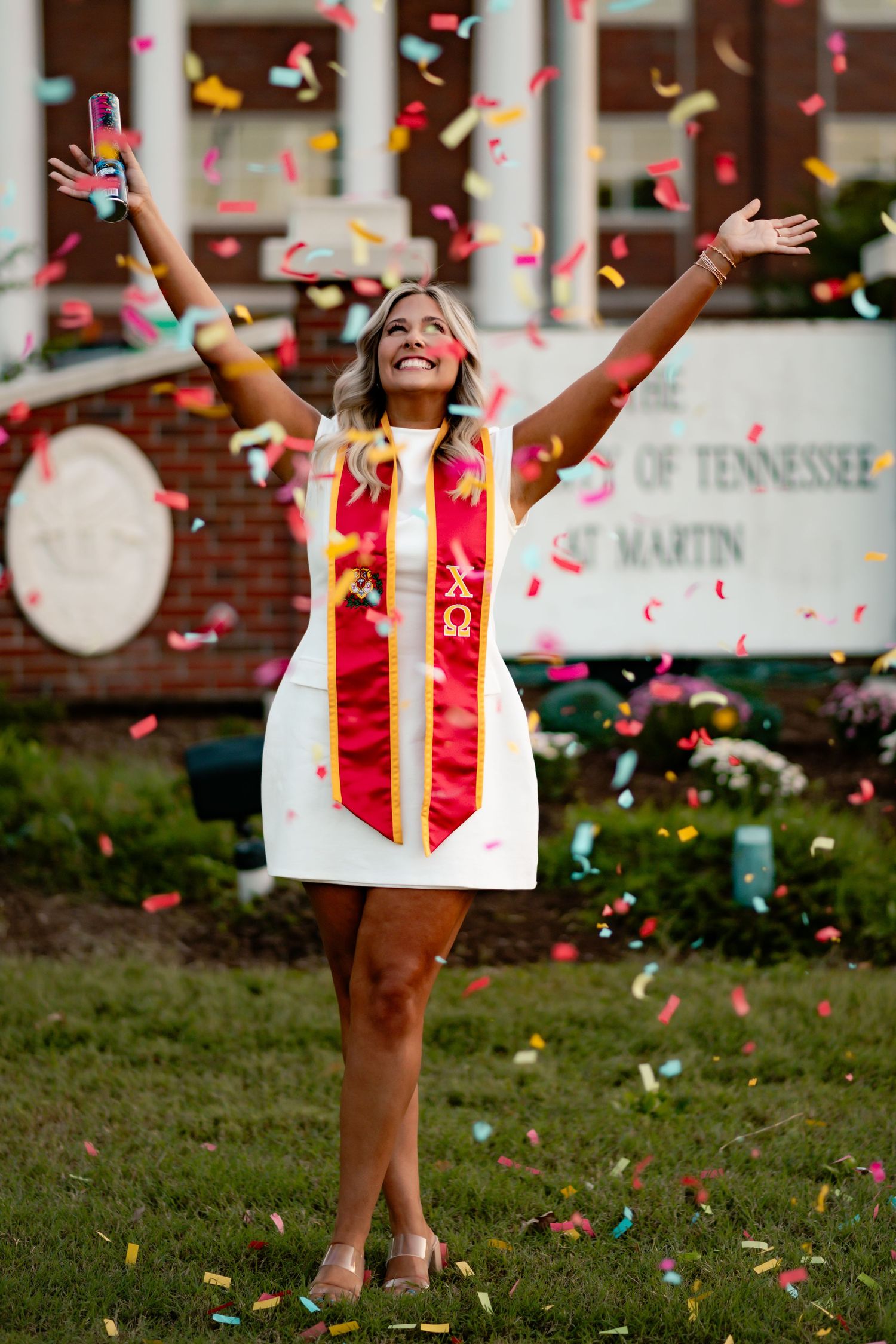Red and pink confetti falls over graduates celebrating outside university building.