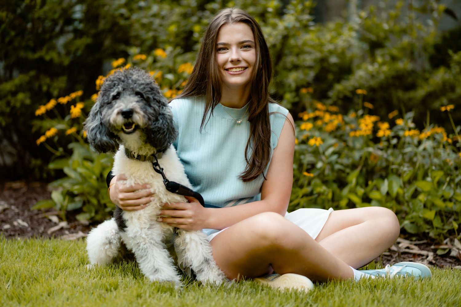 Person sitting on grass outdoors with their dog enjoying a sunny autumn day with yellow flowers in background.