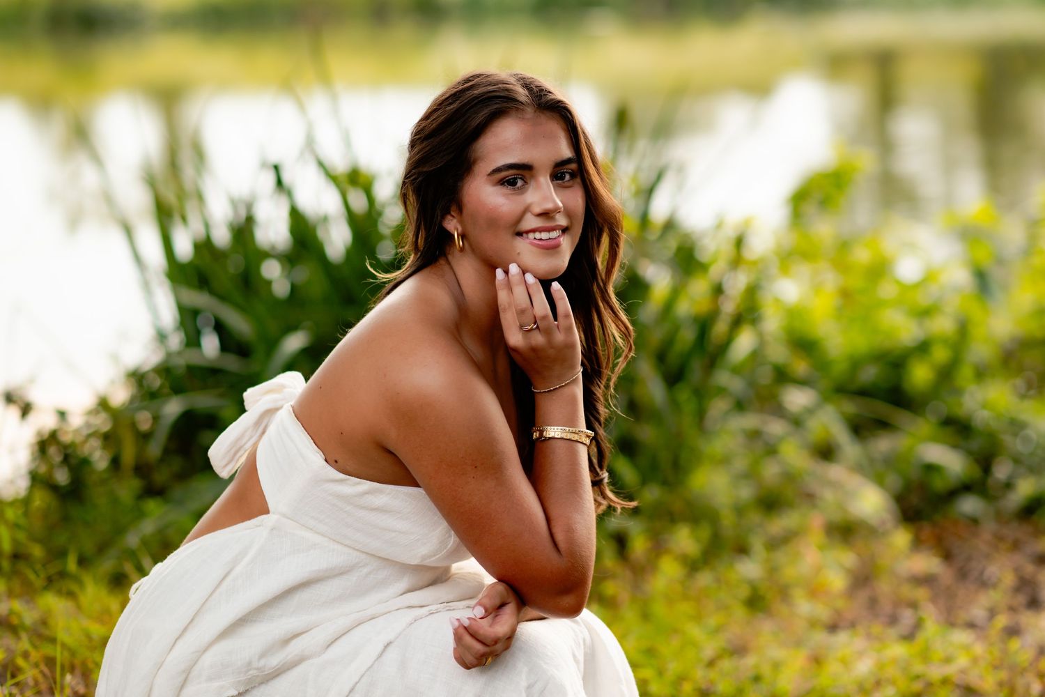 A person in a white dress poses outdoors against a sunlit natural background with greenery and flowers.