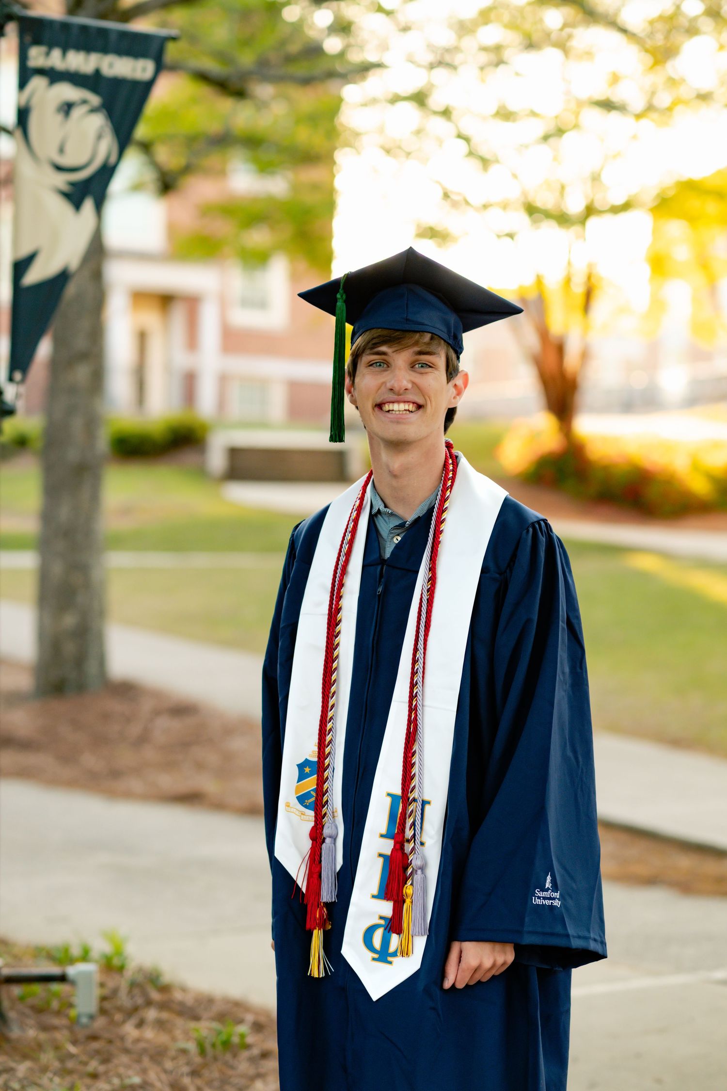 College graduate wearing navy blue regalia and decorated stole stands outdoors during ceremony.