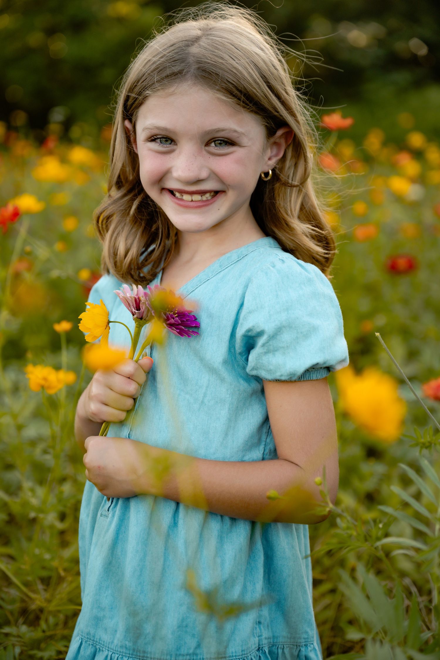 Young child in light blue dress holding wildflowers while standing in a vibrant garden of yellow and red blooms.