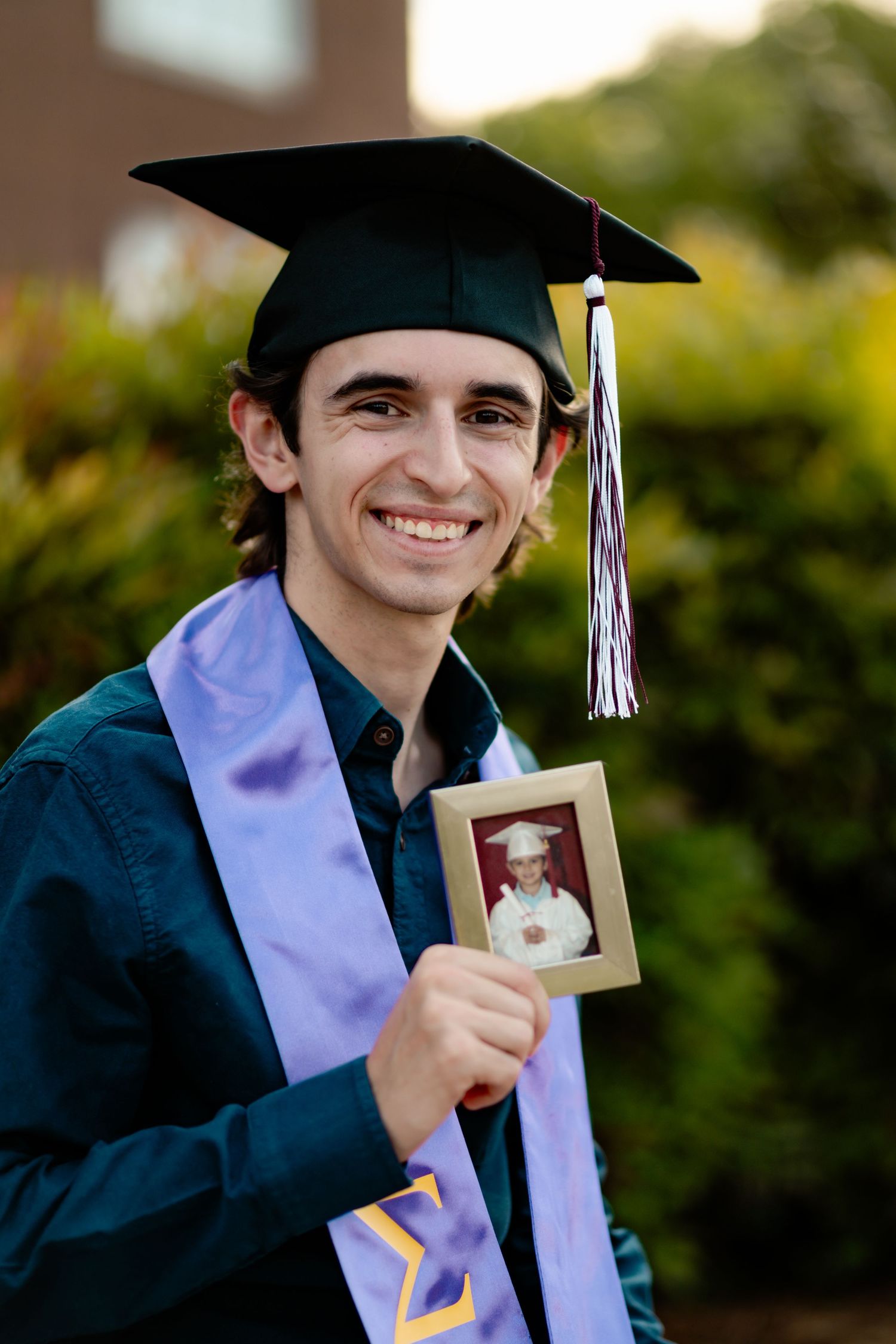 Graduate wearing cap and gown proudly displays photo while smiling during outdoor graduation ceremony.