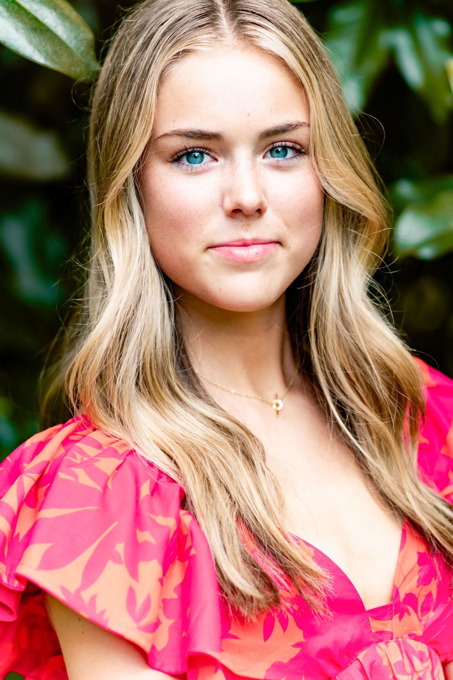 Portrait of someone in a bright pink floral top with wavy blonde hair and blue eyes against a green foliage background.