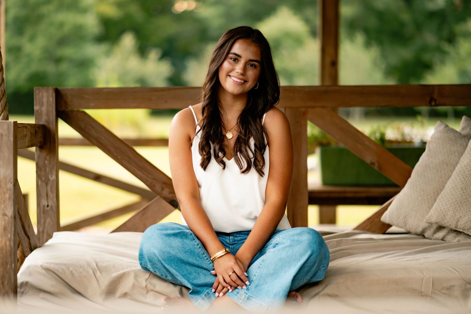 A person sits cross-legged and relaxed on a cushioned outdoor porch bench in a white tank top and blue jeans.
