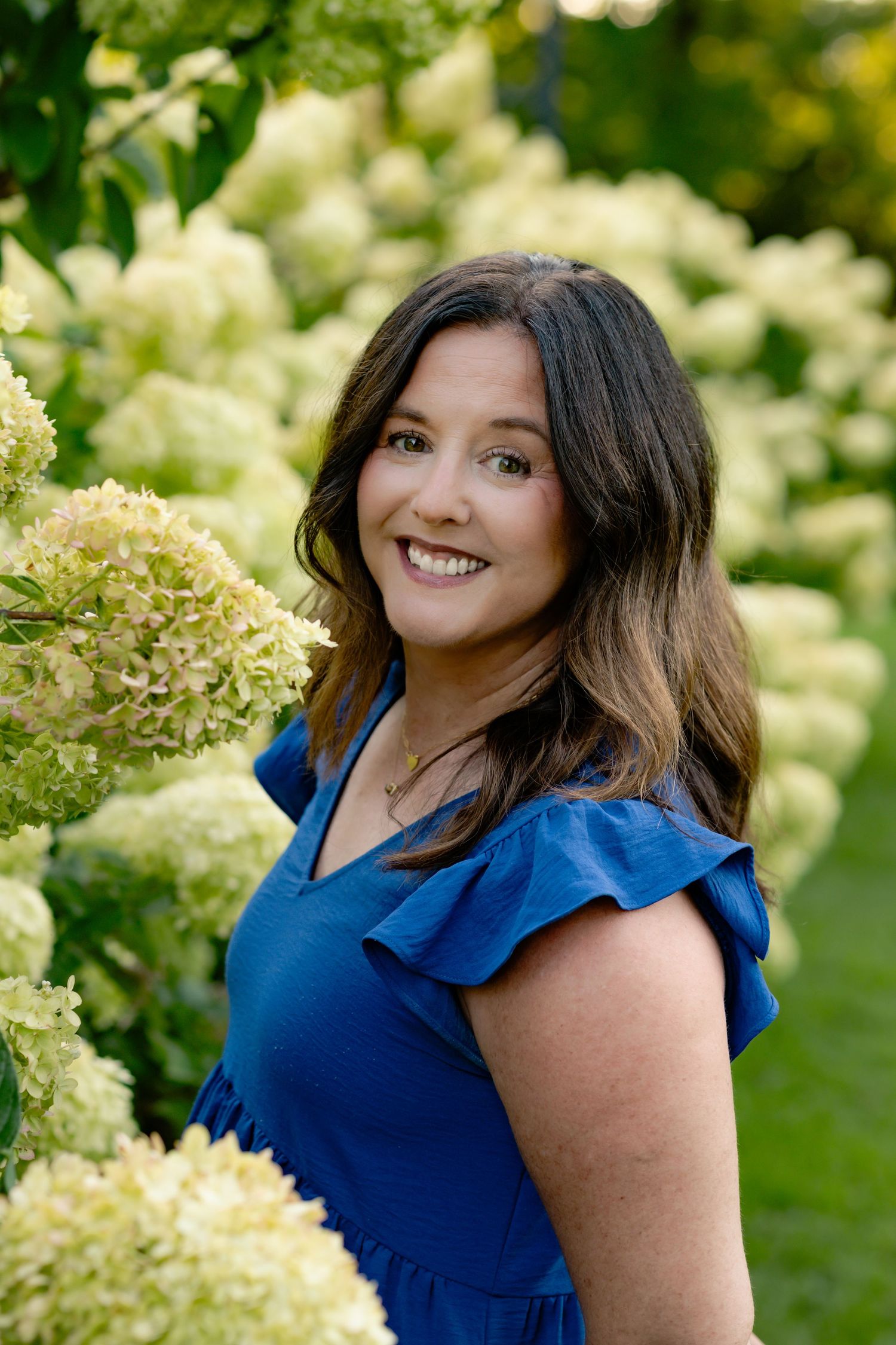 A person in a navy blue ruffled top poses outdoors against a backdrop of white flowering bushes and greenery.