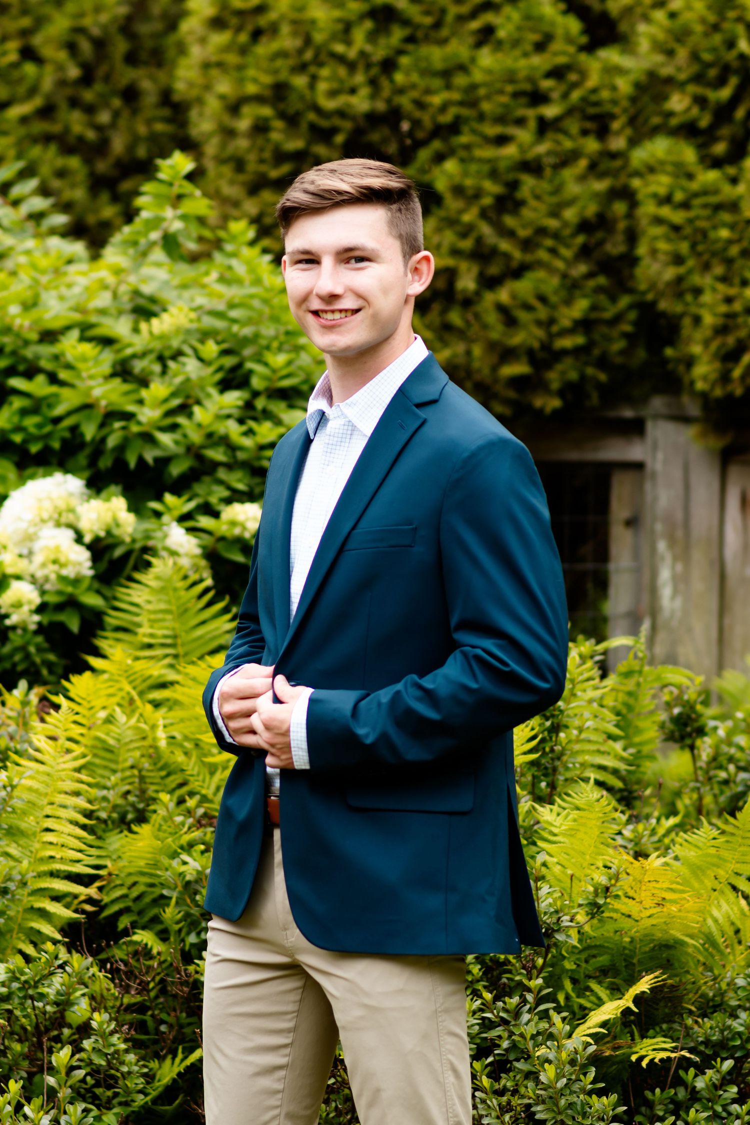 Someone wearing a navy blazer and khaki pants poses for a photo outdoors against lush green ferns and foliage.