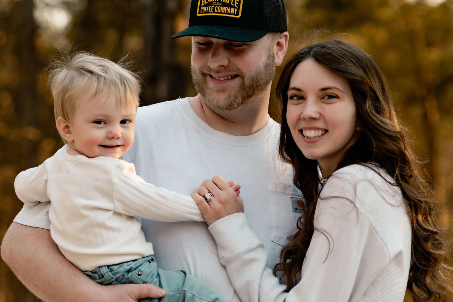 Family of three in coordinating white outfits smiling together during outdoor photo session.